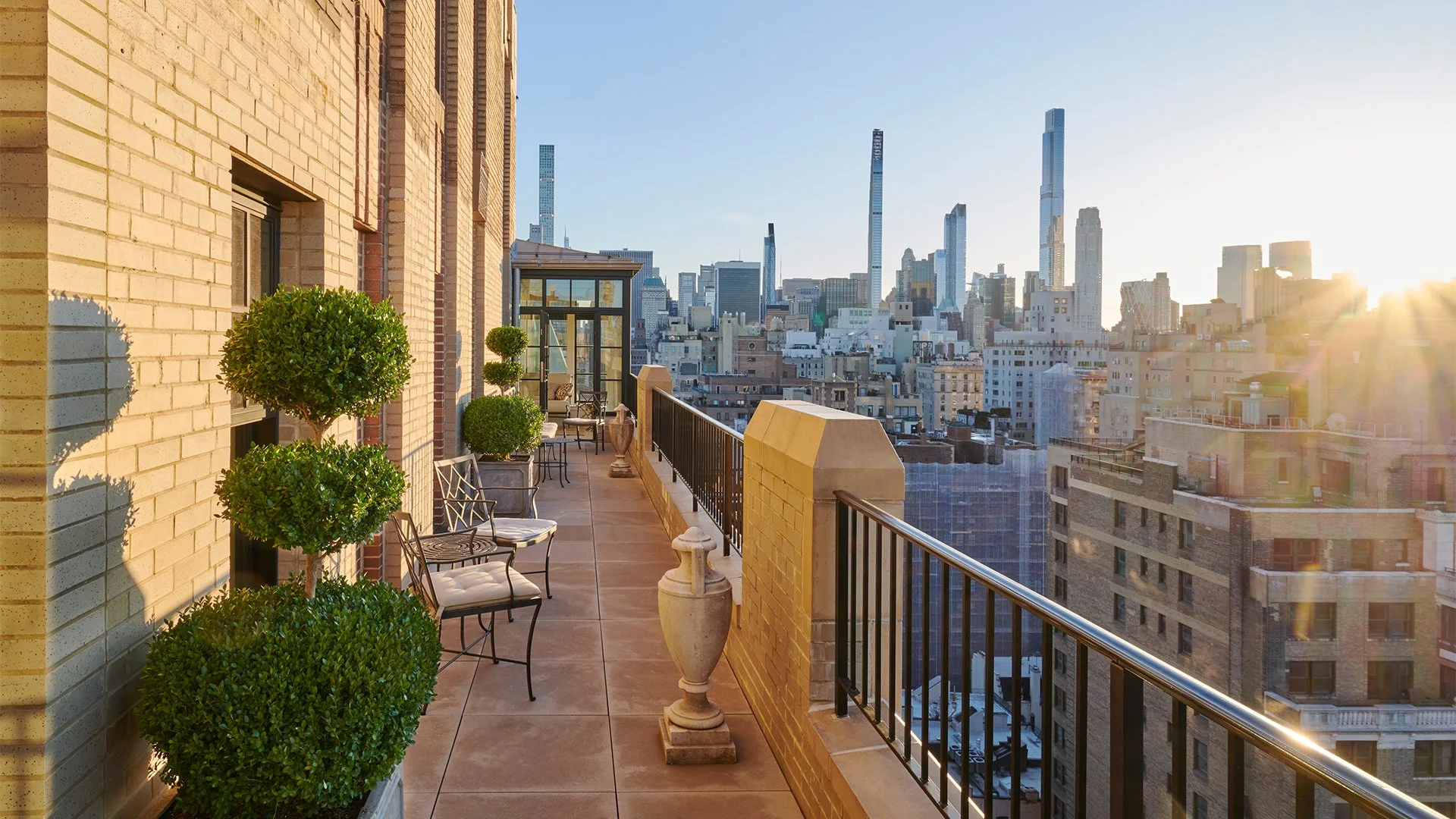 Sunlit balcony with potted topiary trees, chairs, and decorative urns overlooking a city skyline with tall buildings at sunset.