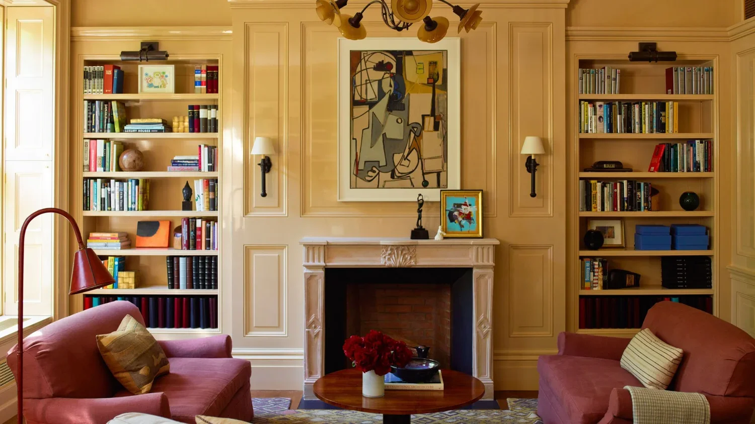 Living room with a fireplace, built-in bookshelves filled with books and decorative items, two pink sofas, a wooden coffee table with a white vase and red flowers, framed artwork above the fireplace, wall-mounted lamps, and a ceiling light fixture.