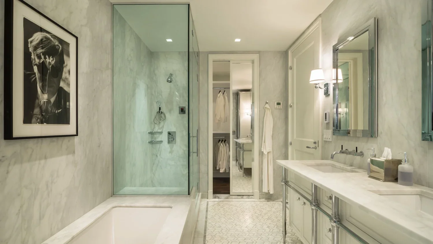 A luxurious bathroom with a marble finish, featuring a shower with glass walls, a bathtub, a vanity with a sink, mirrors, and wall-mounted lighting. There's a framed black-and-white portrait on the wall, and a closet area with hanging robes visible t