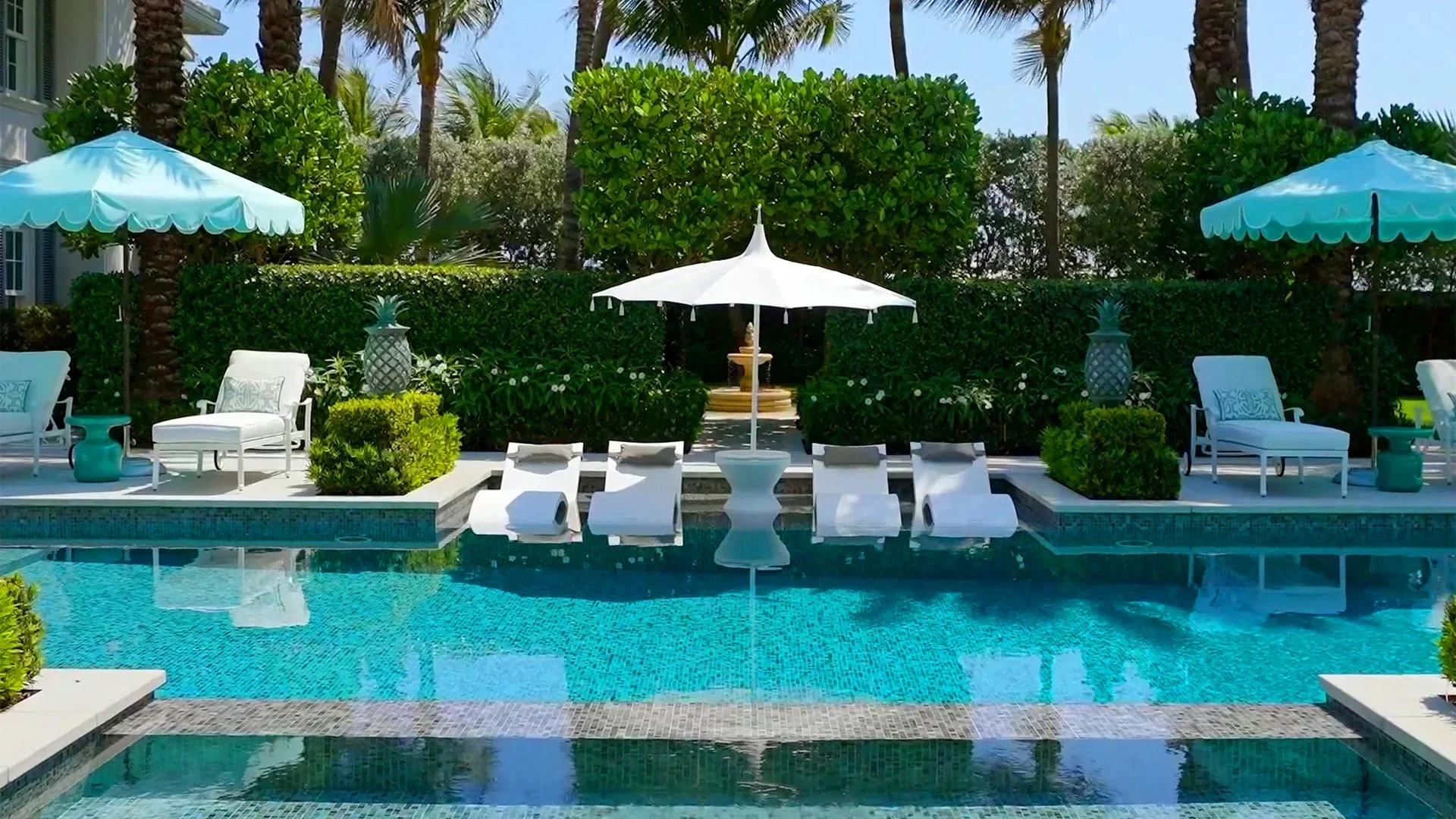 Luxury swimming pool area with white lounge chairs, umbrellas, lush tropical plants, and a decorative fountain, set against a backdrop of green hedges and palm trees.
