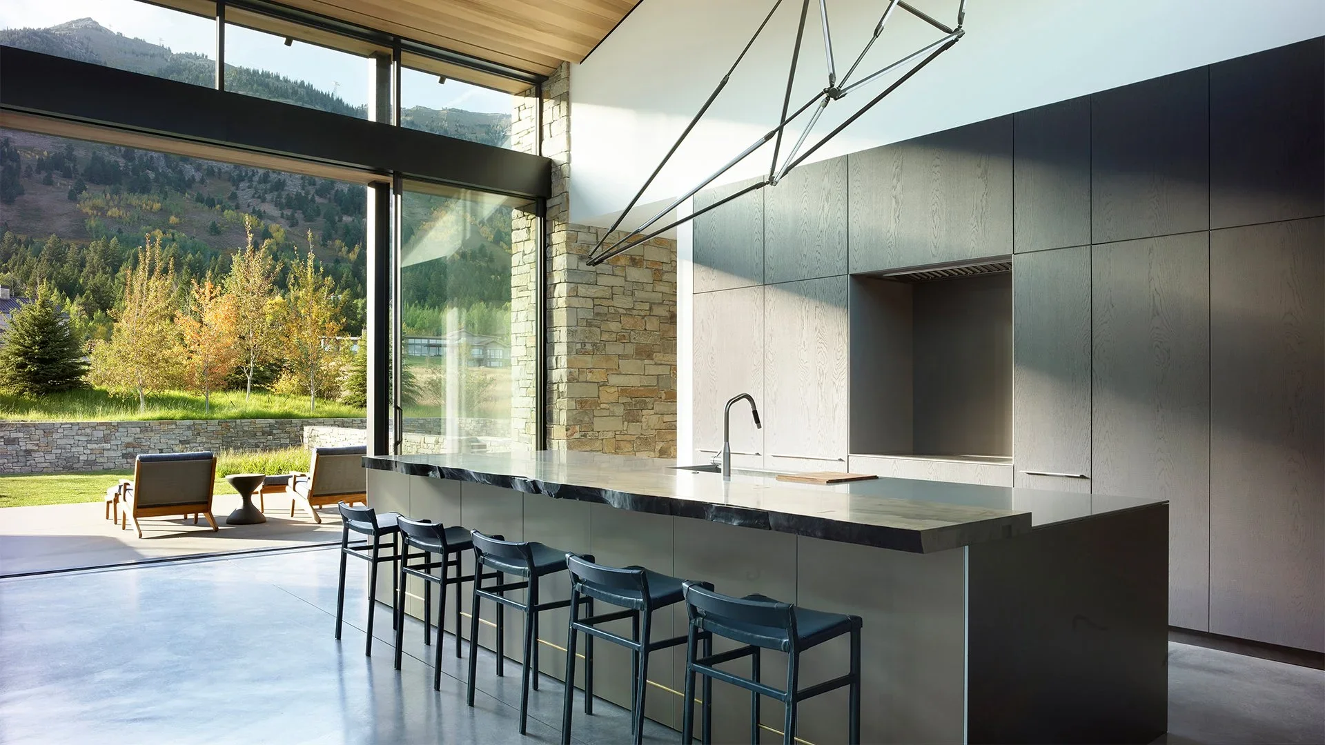 Modern kitchen with a large marble island, black stools, and a view of a garden with trees and mountains outside.