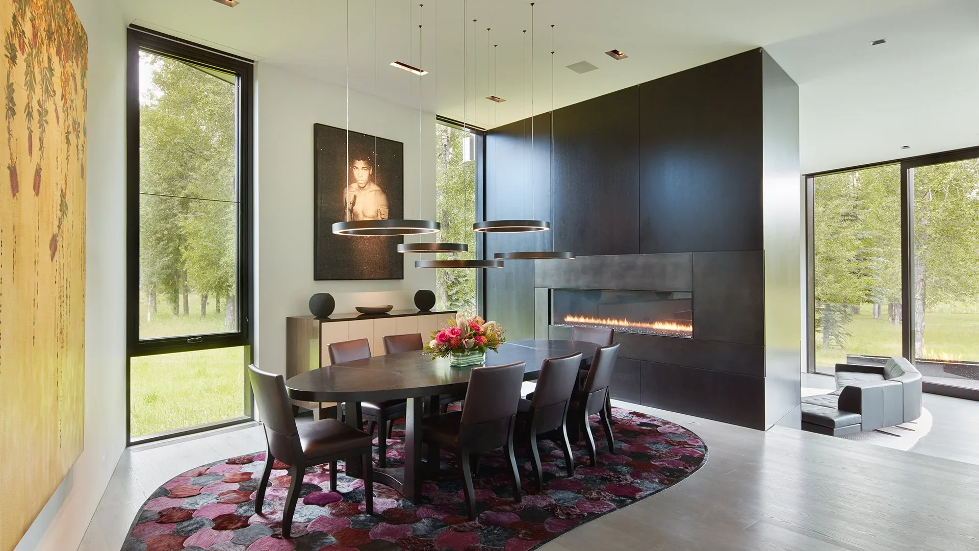 Modern dining room with a large oval table, six chairs, floral centerpiece, fireplace, large windows with views of green trees, and contemporary artwork on the wall.