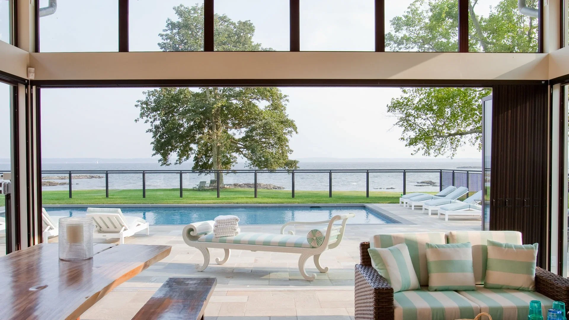 Interior view of a living room opening to an outdoor pool area with a scenic ocean view, including trees, lounge chairs, and a grassy lawn.
