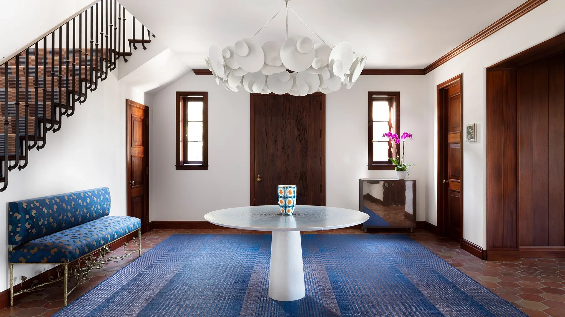 Modern living room with white walls, wooden door and trim, a blue patterned bench, a round white table with a decorative vase, a blue rug, and a unique white chandelier hanging from the ceiling.
