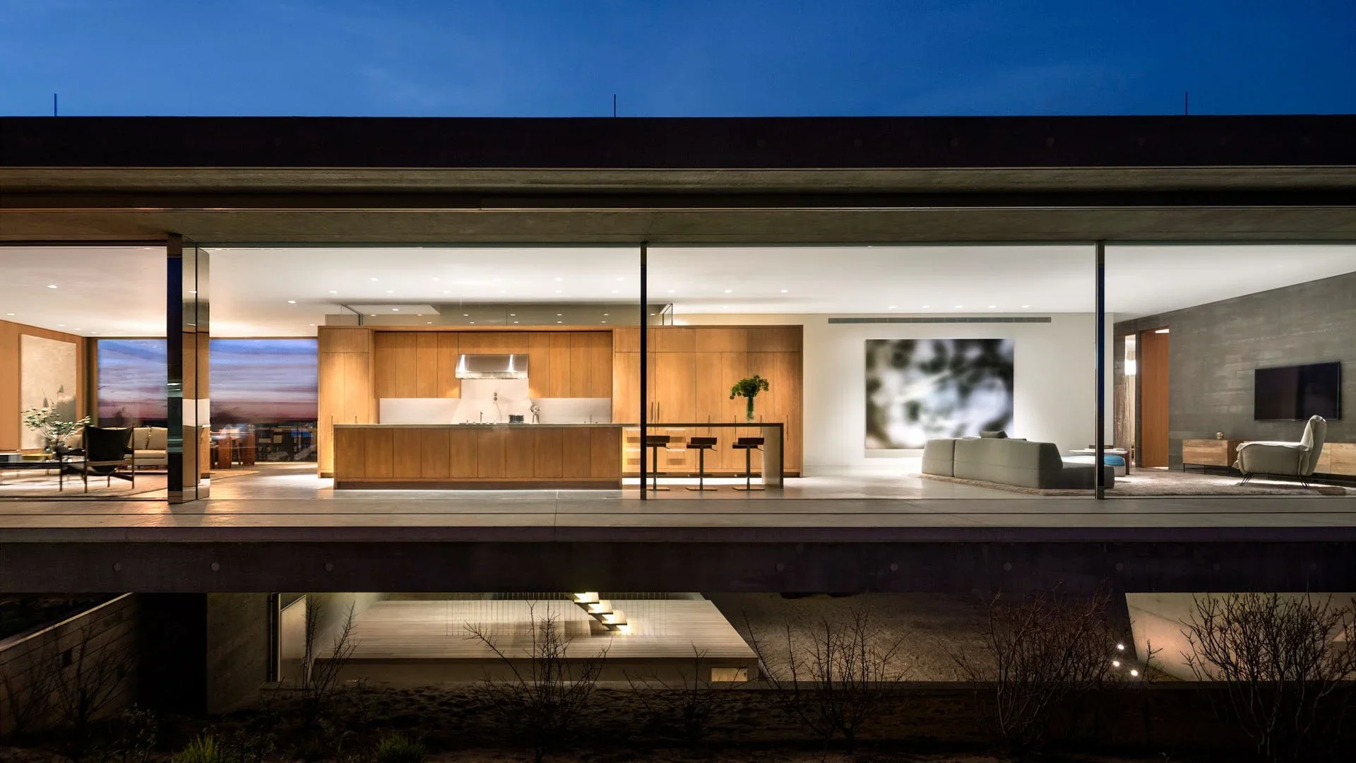 Modern house interior with open kitchen and living room, seen through large glass windows at night.