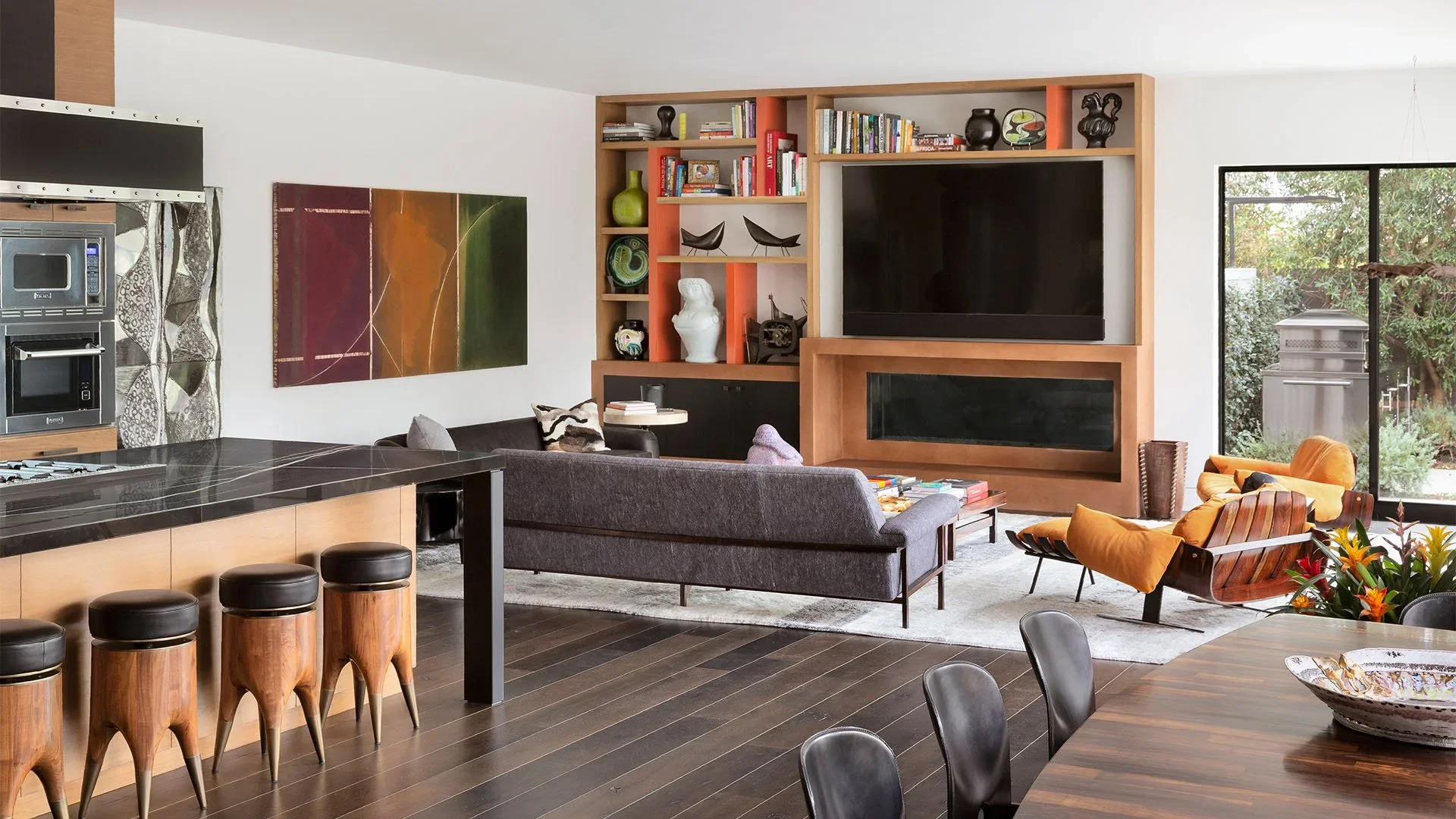 A modern living room with a gray sofa, a yellow armchair, a large flat-screen TV, a built-in fireplace, and a bookshelf filled with books and decorative items. There is a large window showing outdoor greenery, a dining table with black chairs, and a 