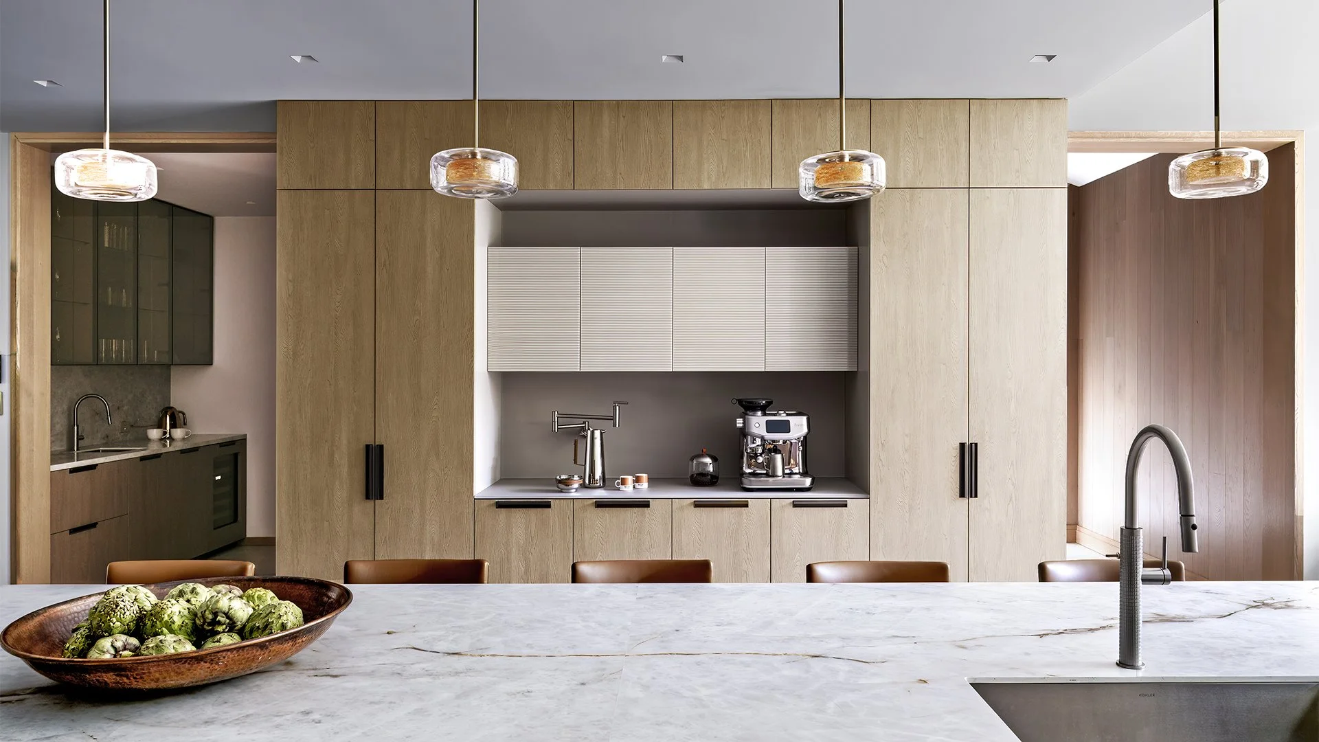 Modern kitchen with beige and gray cabinetry, hanging pendant lights, a marble kitchen island with artichokes in a bowl, and a coffee machine with cups on the countertop.