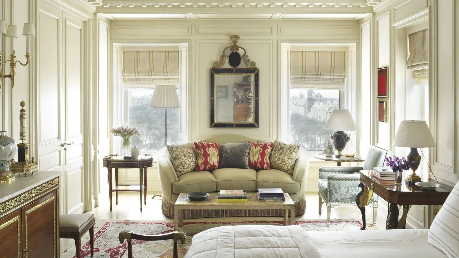 Elegant living room with large windows, cream-colored walls, and classic furniture including a sofa with decorative pillows, armchairs, tables with lamps, and various decorative items.