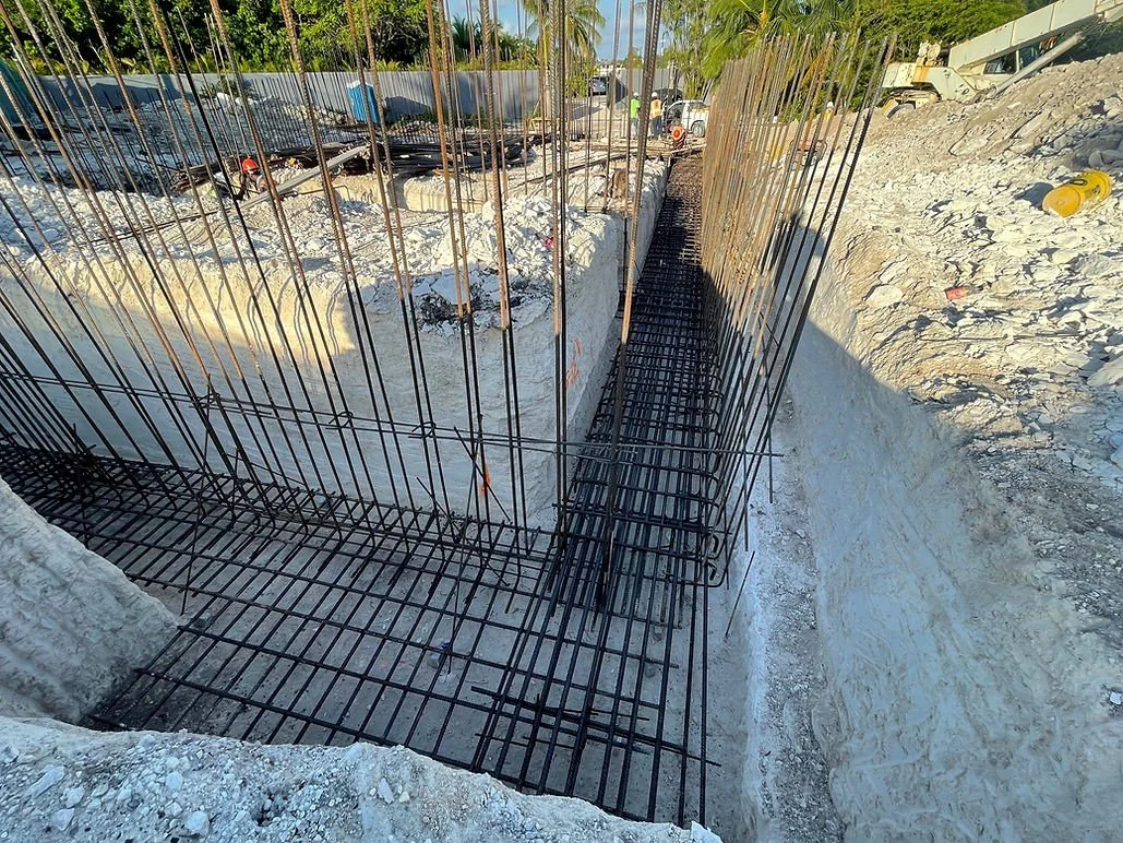 Construction site with rebar reinforcement extending into a deep trench, preparing for concrete pouring, surrounded by rocky soil and construction equipment in the background.
