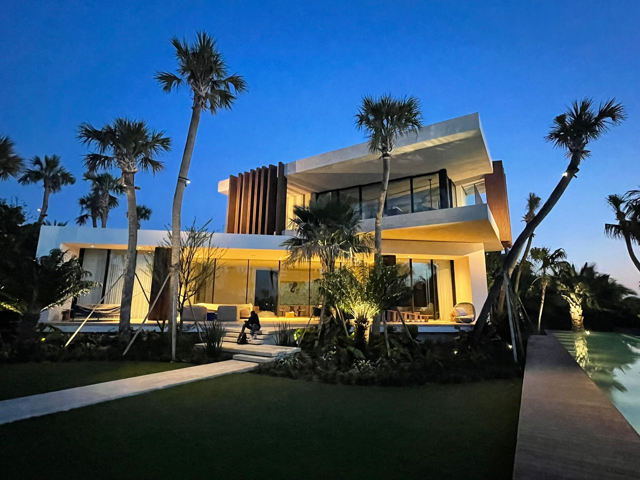 Modern luxury house with large glass windows, surrounded by palm trees and landscaped yard, during twilight.