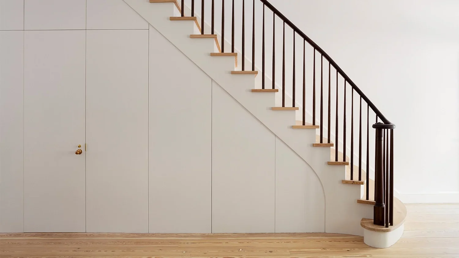 Interior view of a staircase with wooden steps and black railing, adjacent to a white wall and floor with wooden planks.