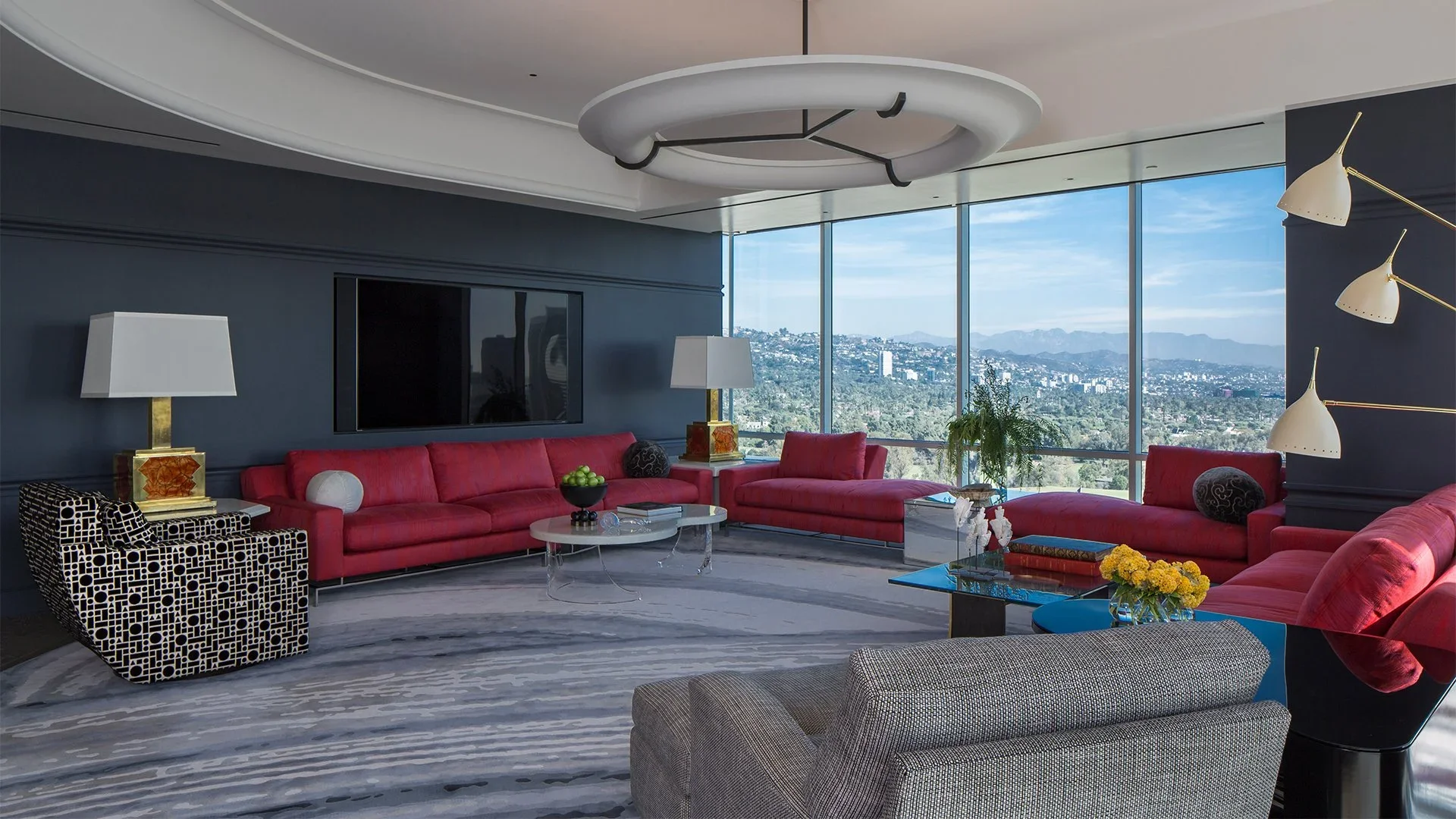 Modern living room with large windows showcasing cityscape view, red sofas, a patterned armchair, and contemporary decor including lamps, plants, and coffee tables.