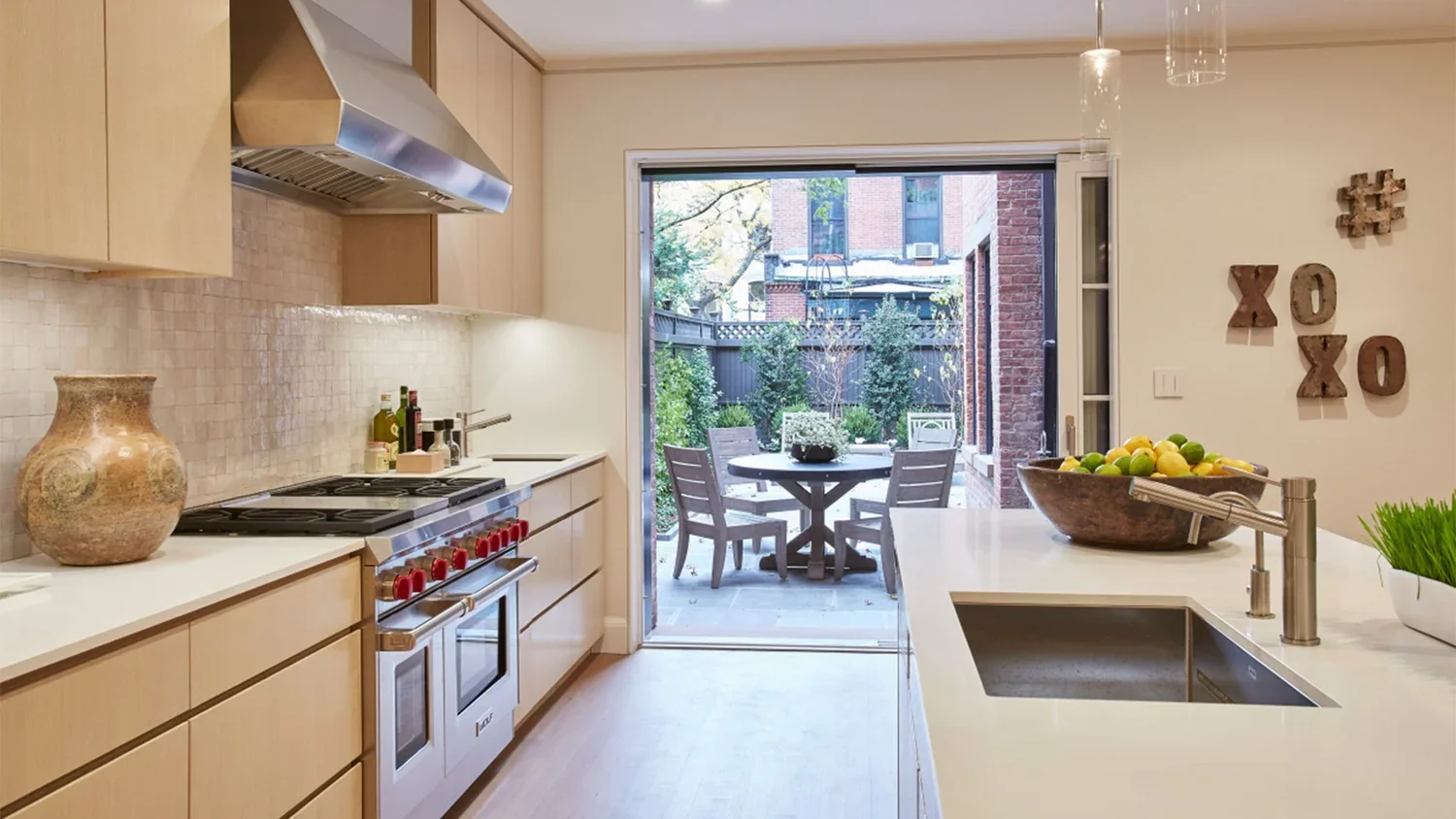 Modern kitchen with beige cabinets, white countertops, and a view of an outdoor patio with a round table and four chairs surrounded by green plants.