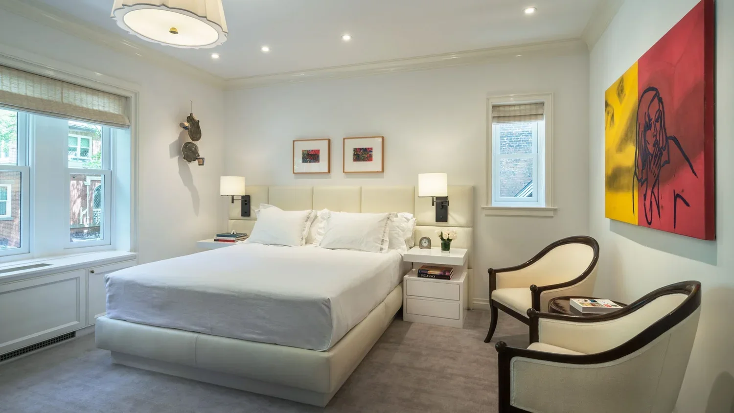 A bedroom with a large bed, white bedding, and two bedside tables with lamps. There are two windows, framed artwork above the bed, and a colorful abstract art piece on the wall. Two white armchairs are placed near the corner, and a ceiling light fixt