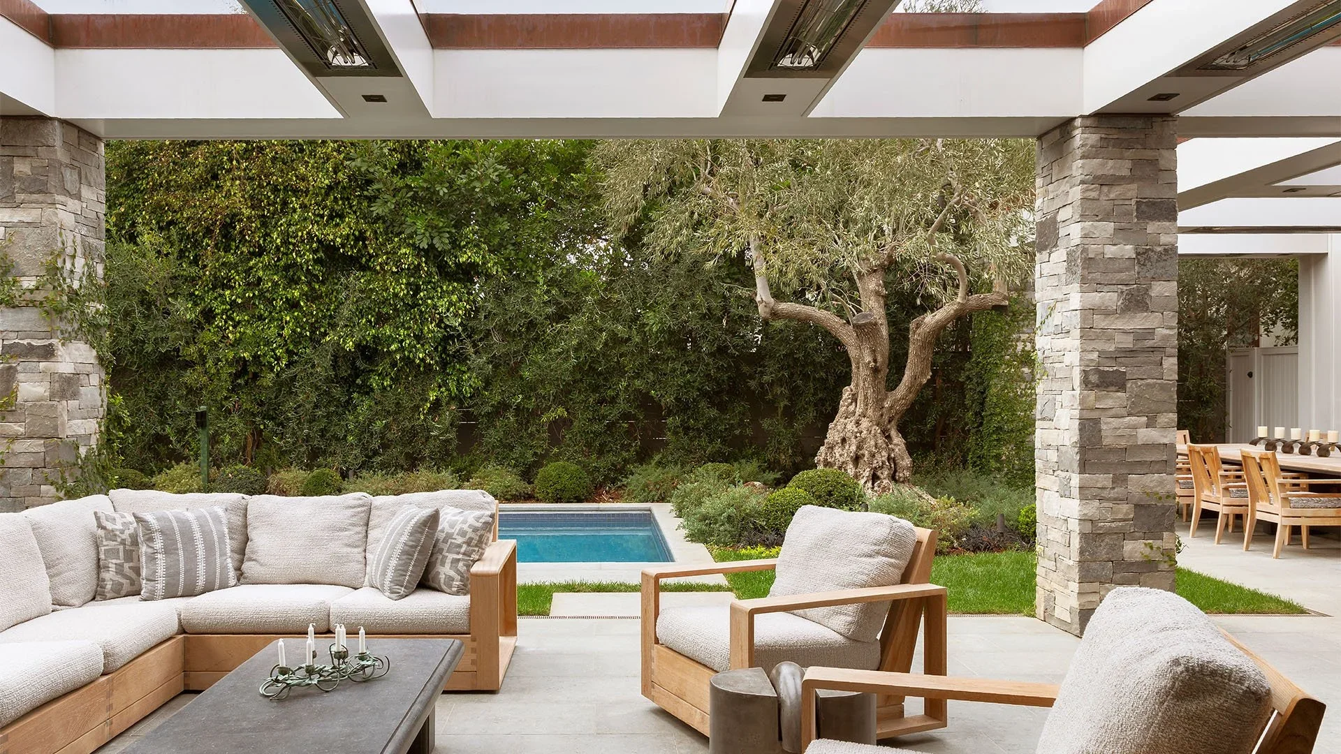 Outdoor patio with beige cushioned wicker furniture, a wooden dining table with chairs, a small pool, and a large twisted tree in a backyard with lush greenery and stone pillars.