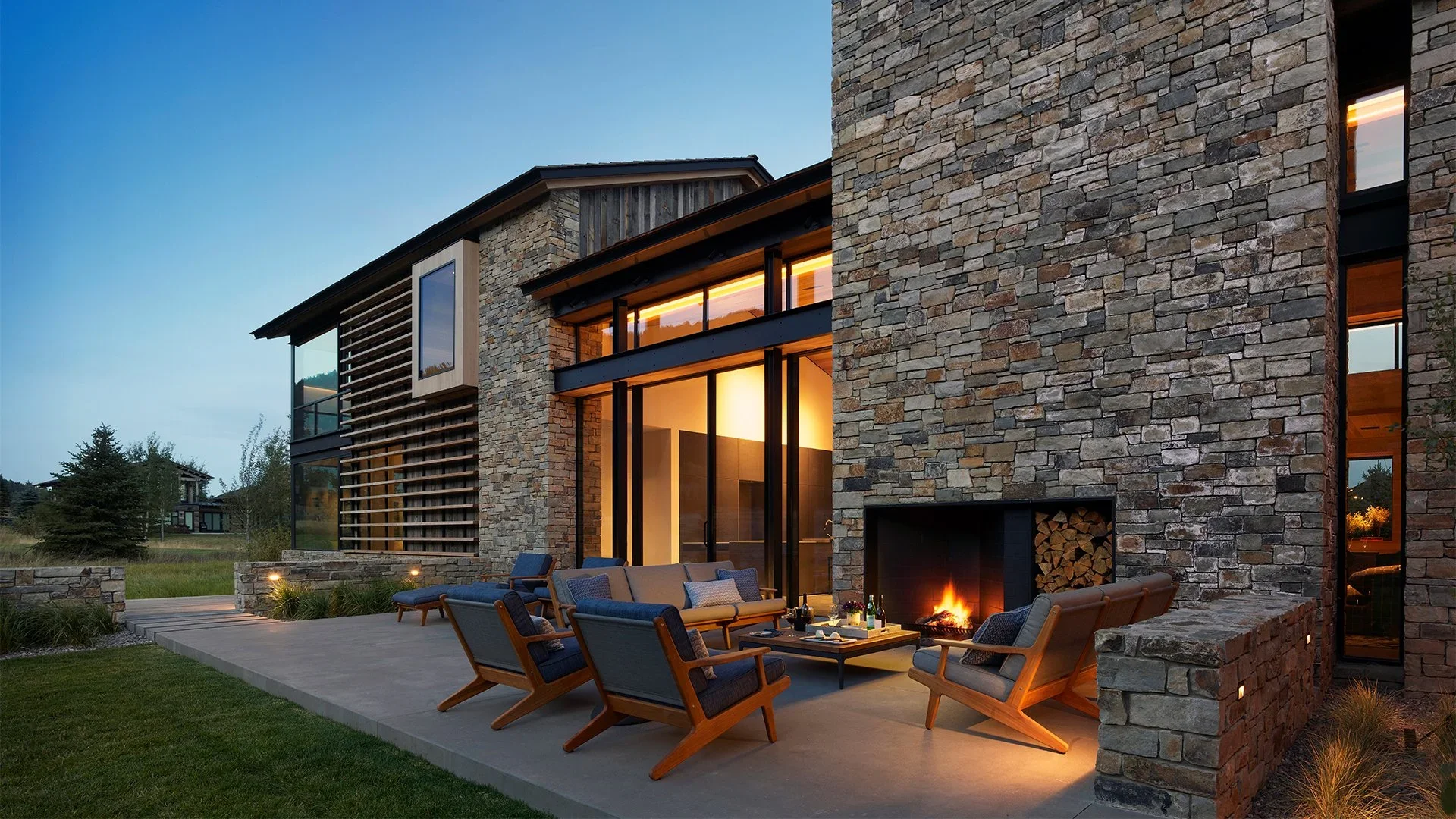 Modern house with stone walls and large glass windows at dusk, outdoor seating area with chairs, sofa, and fireplace.