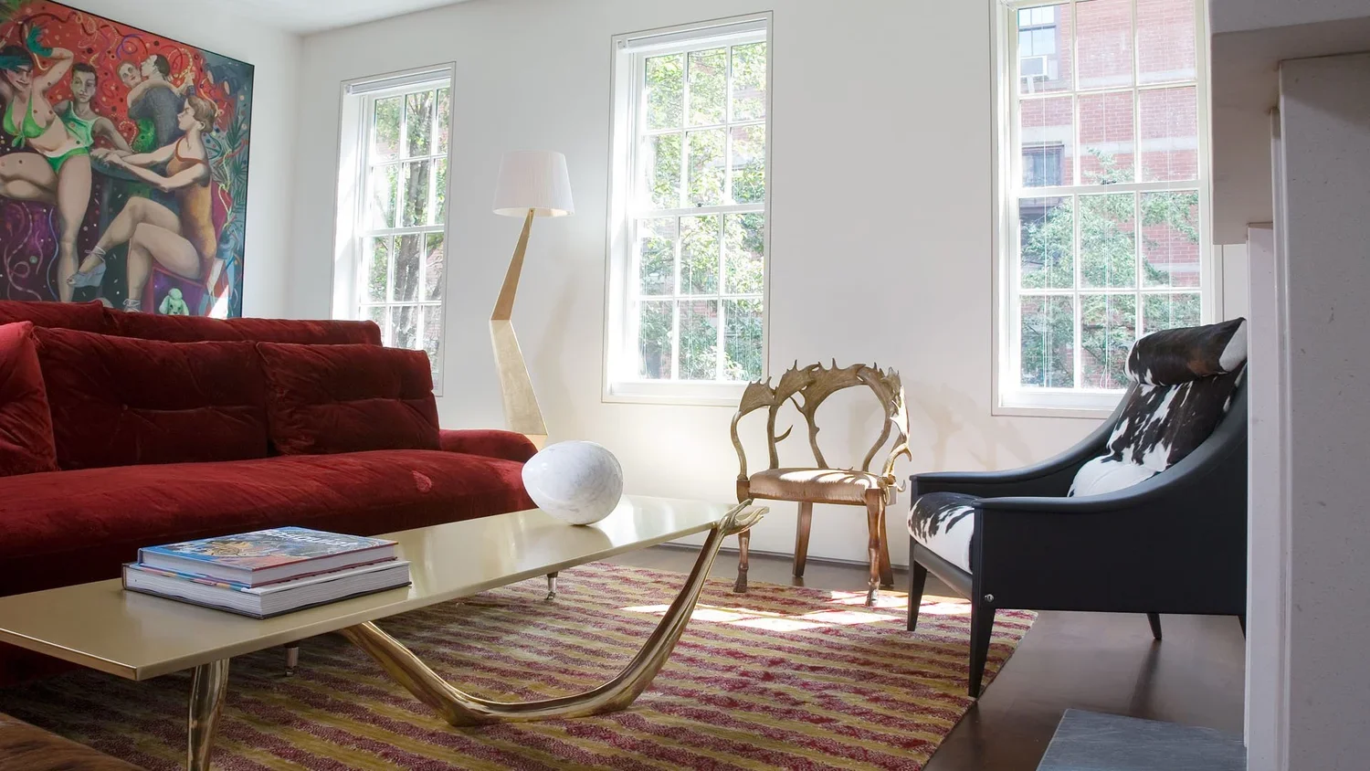 Living room with a red velvet sofa, a modern beige floor lamp, a decorative wooden chair, a black and white cowhide armchair, a striped area rug, and artwork on the wall with large windows letting in natural light.