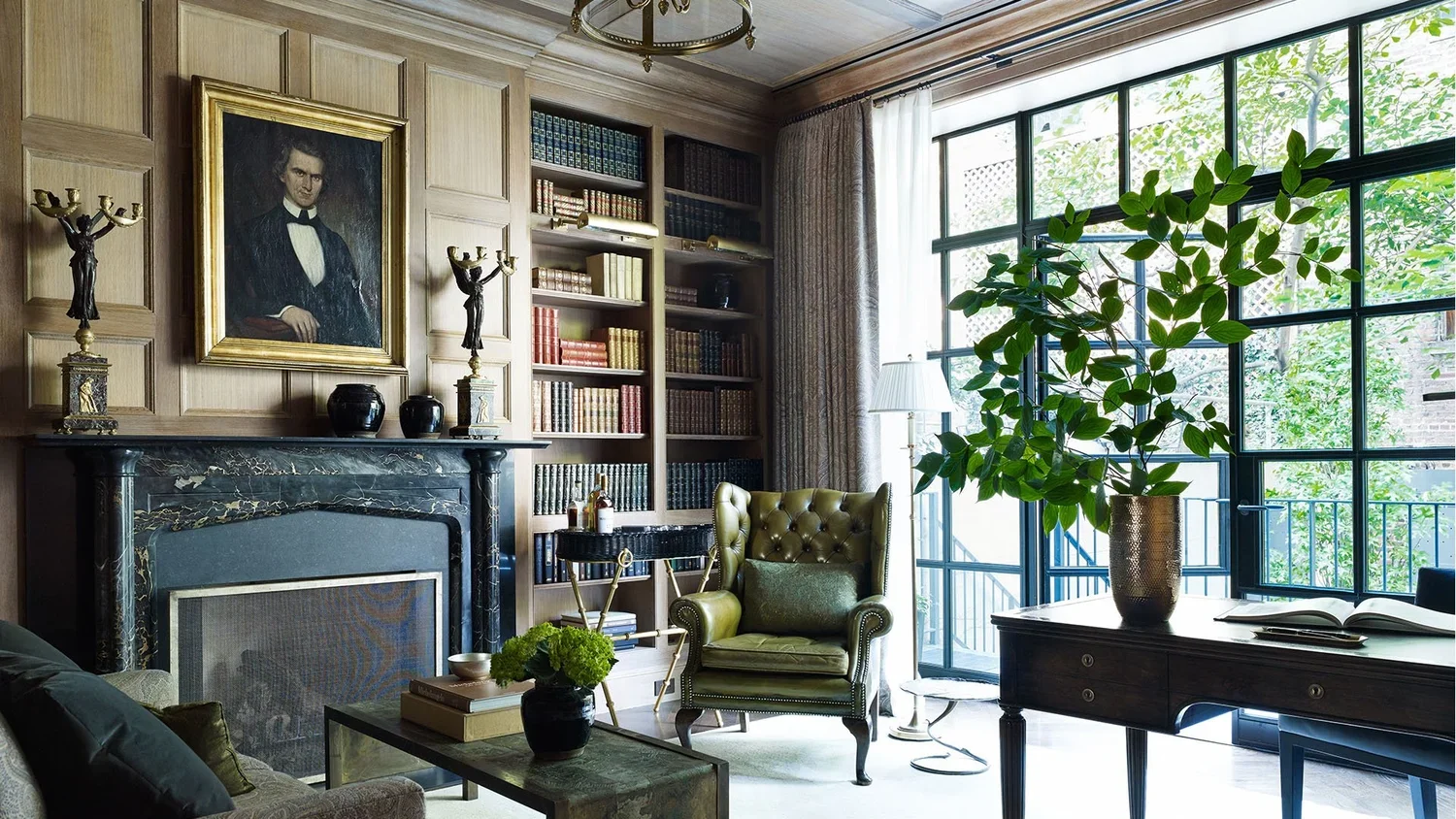 Interior of a living room with a fireplace, a portrait above the fireplace, bookshelves filled with books, a green leather armchair, a large window with curtains, a black table with a potted plant, and a side table with a lamp and a large vase with g