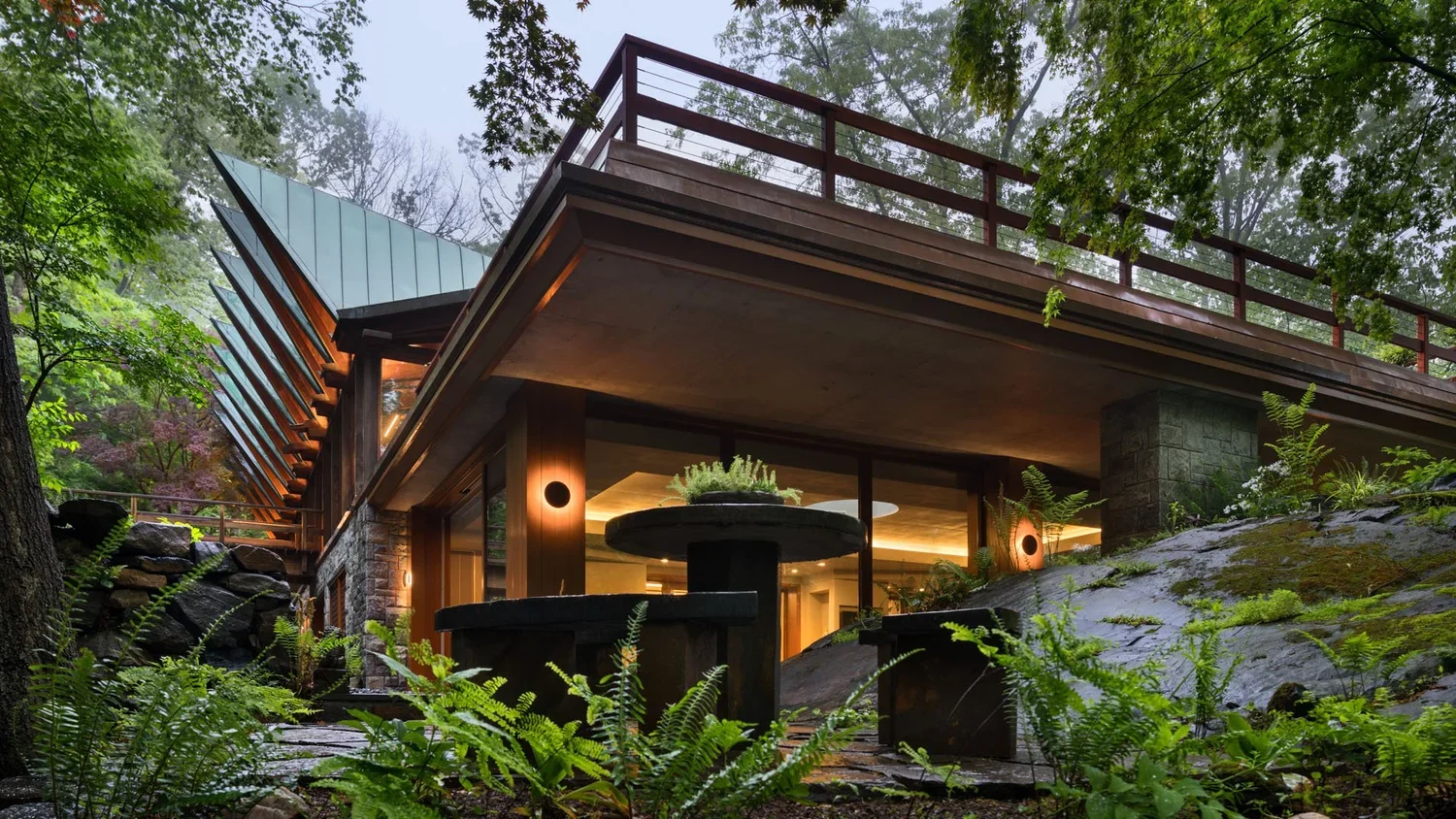Modern house nestled among trees with outdoor seating and large windows, surrounded by lush greenery.