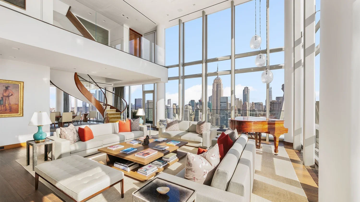 Spacious living room with large windows overlooking New York City skyline, white sofas with colorful cushions, a grand piano, a wooden coffee table with magazines, and a spiral staircase.