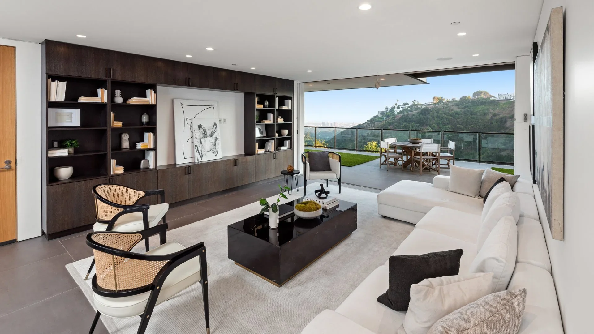 Modern living room with white sectional sofa, black coffee table, armchair, built-in dark wood shelving, large sliding glass door leading to balcony with outdoor dining table, view of hillside and blue sky