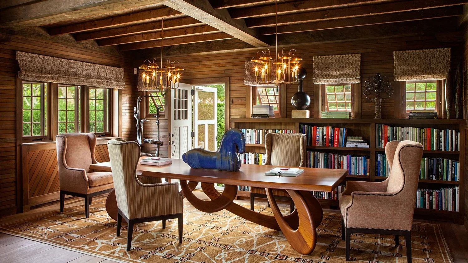 A cozy, rustic interior room with wooden walls, ceiling, and furniture, featuring a large wooden table surrounded by upholstered chairs, a bookshelf filled with books, natural light from multiple windows, decorative chandeliers, and a door opening to