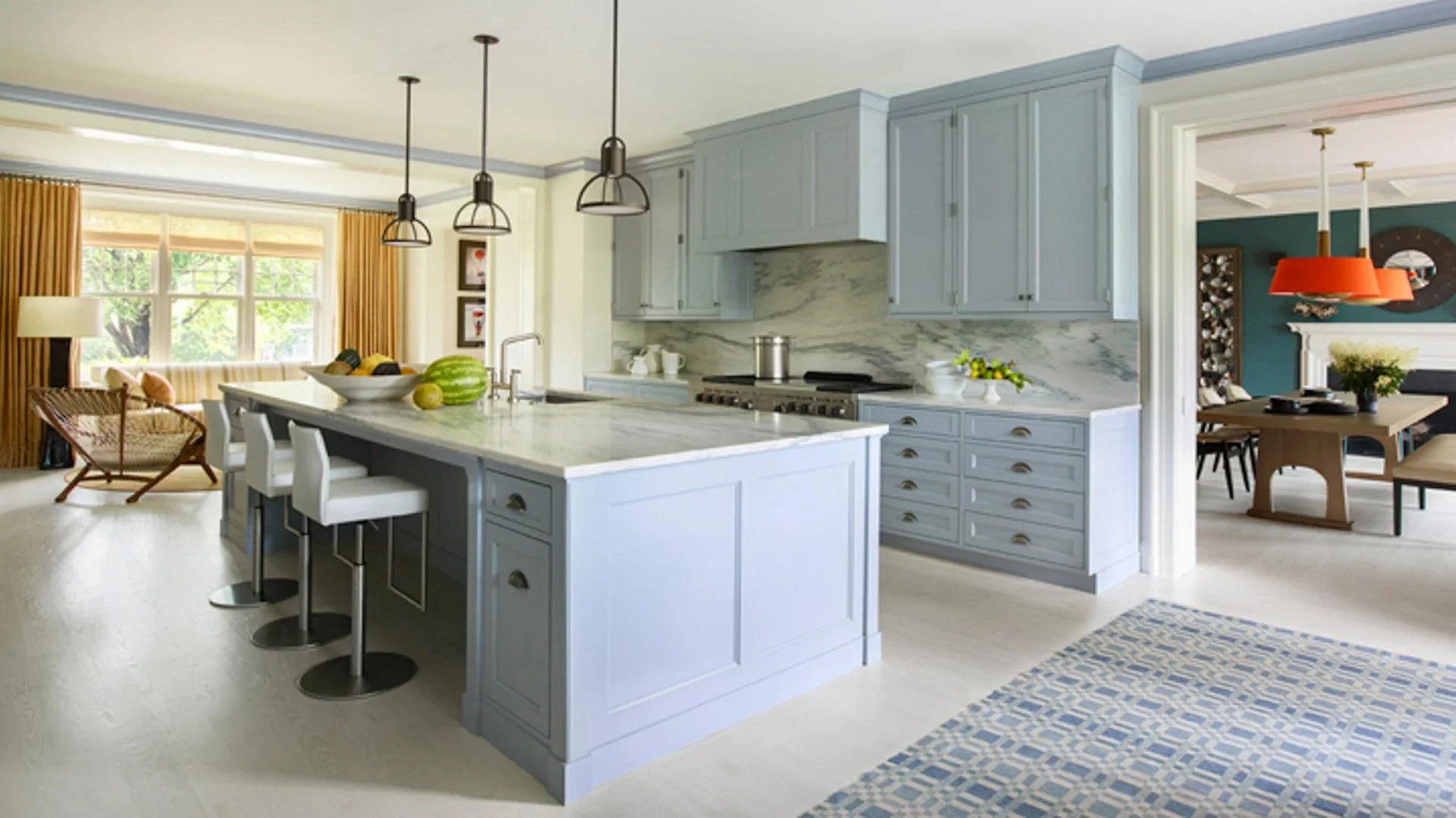 Open-concept kitchen and dining area with light blue cabinetry, white marble countertops, and a kitchen island with three white barstools. Adjacent living space with large windows, yellow curtains, and modern furniture. Dining area with wooden table,