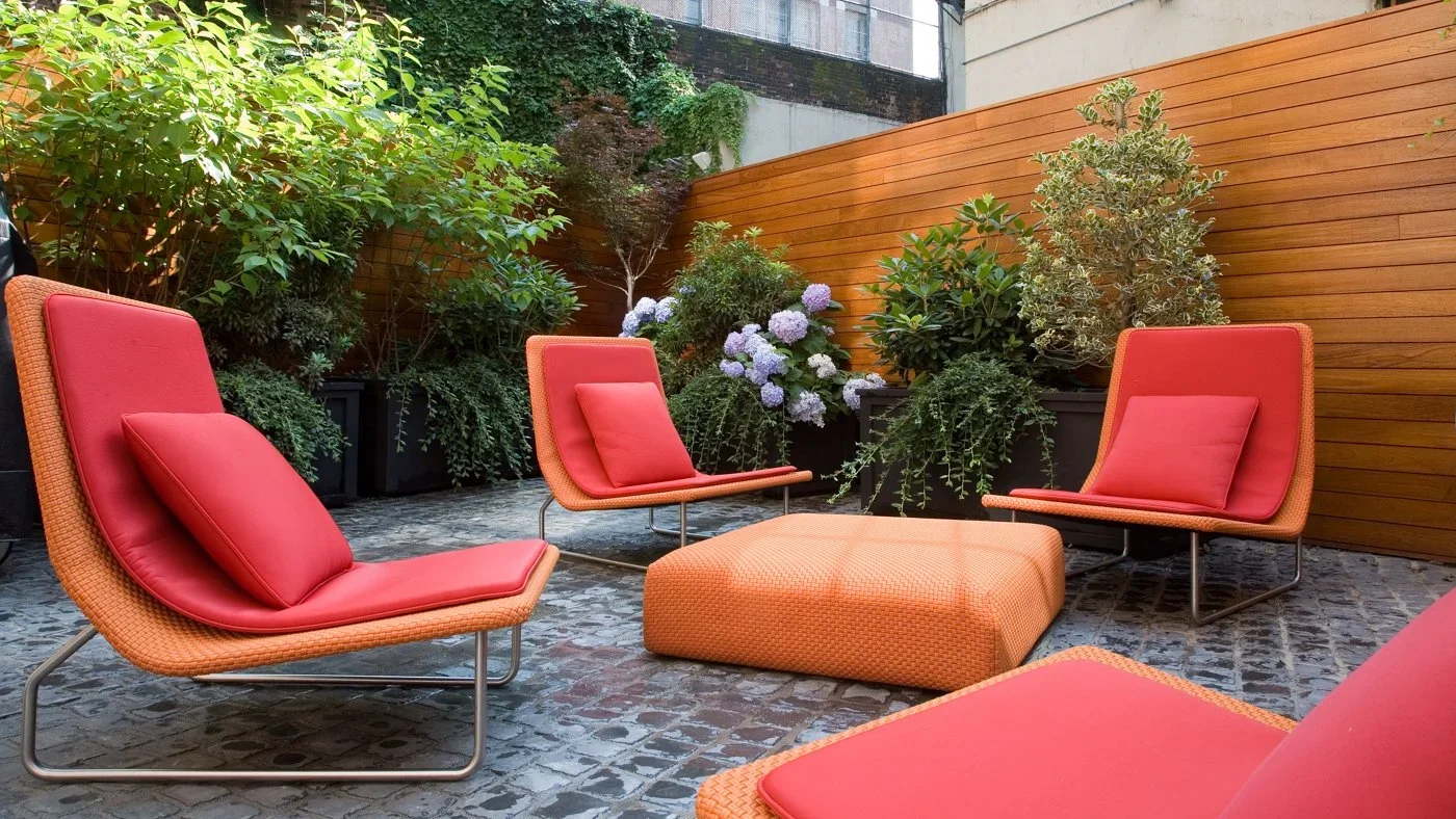 Outdoor patio with four orange and red lounge chairs, an orange ottoman, lush greenery, flowering plants, and a wooden privacy fence.