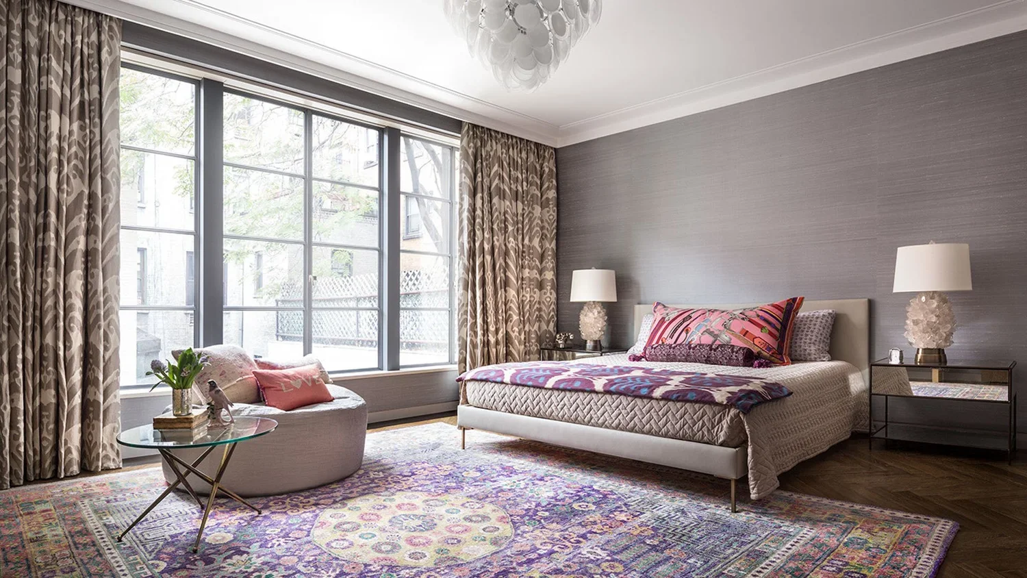 A bedroom featuring a large window with beige patterned curtains, a bed with a pink and purple bedspread, white lamps on black side tables, a round armchair with pink pillows, a glass side table with a vase of flowers, and a colorful Persian rug on d