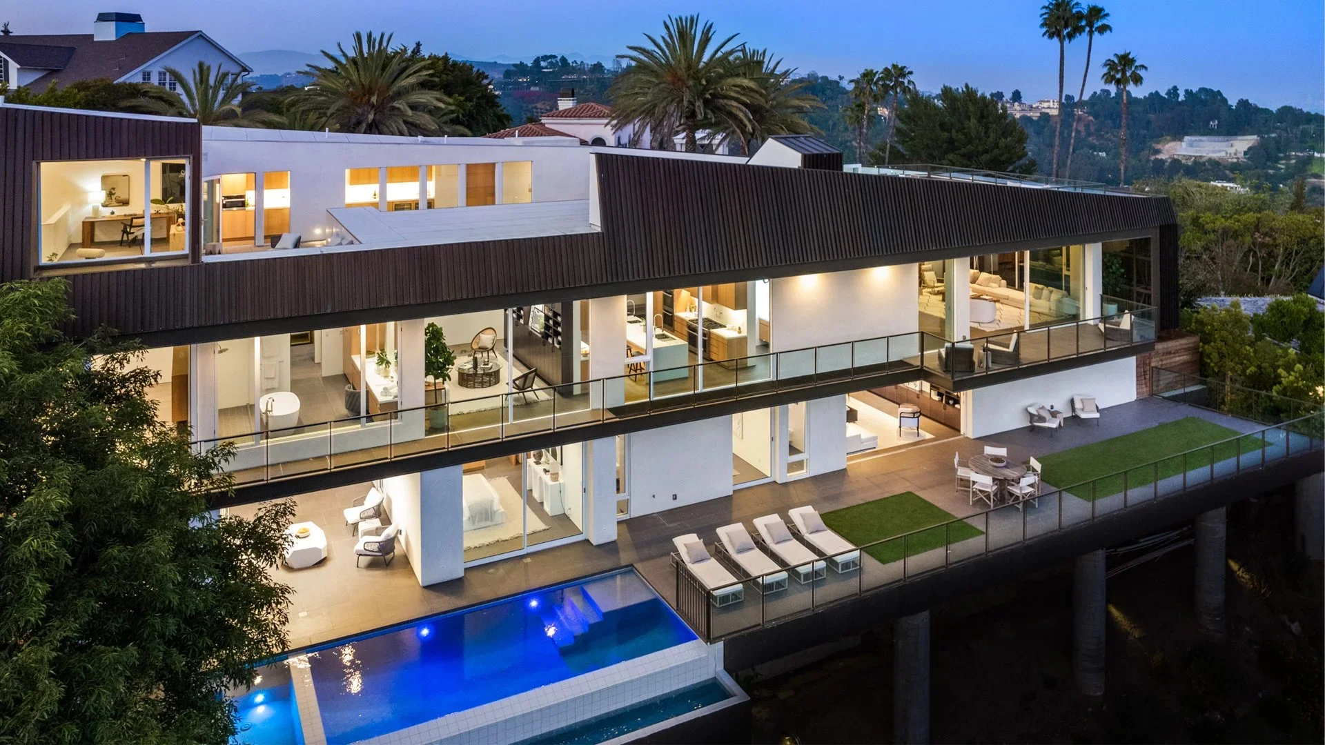 Modern multi-level house with large glass windows, an outdoor swimming pool, and a spacious balcony, set amidst lush greenery and palm trees at dusk.