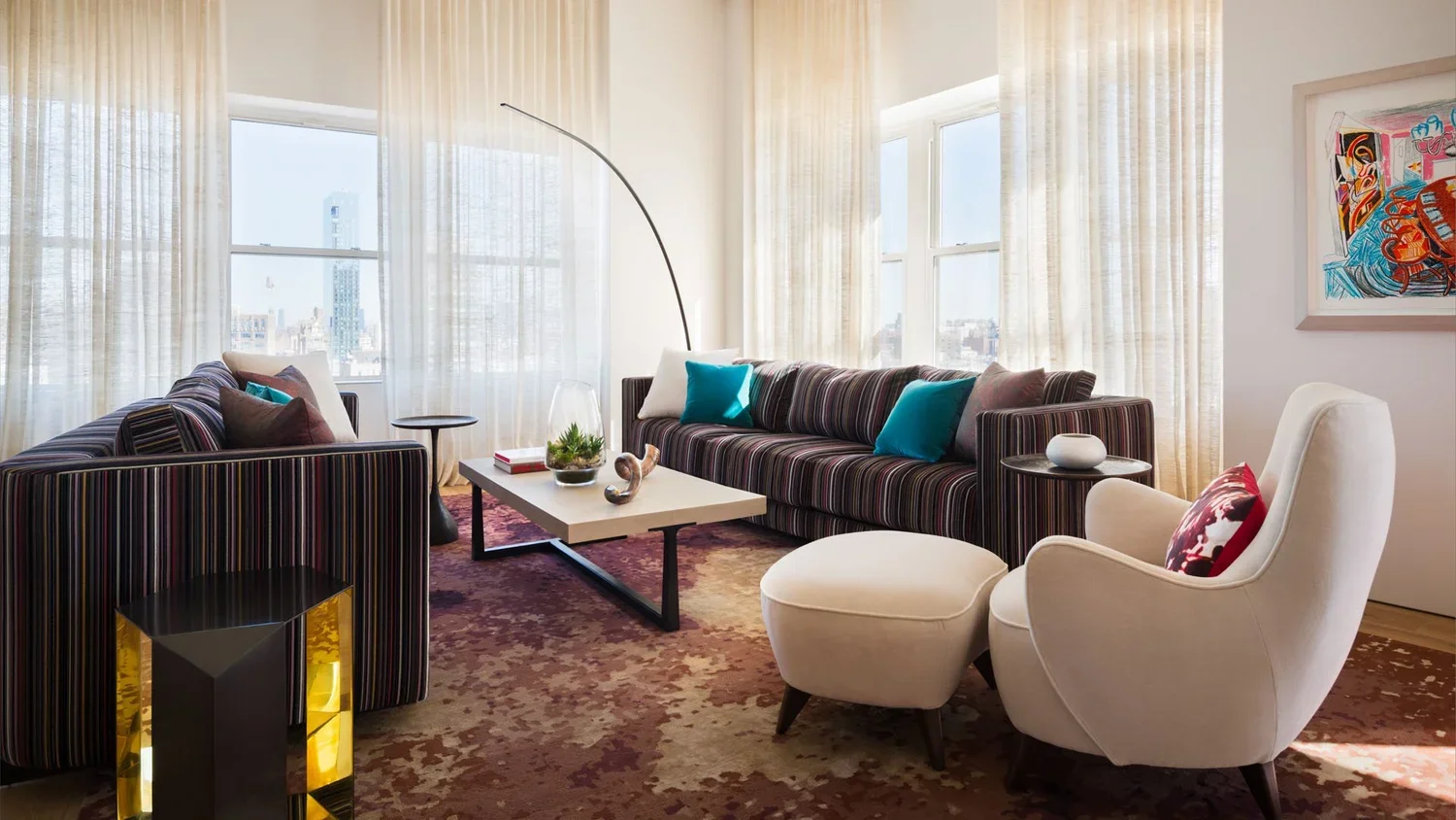Modern living room with two large striped sofas, a white armchair with an ottoman, a coffee table, and large windows with sheer curtains, cityscape view in background.