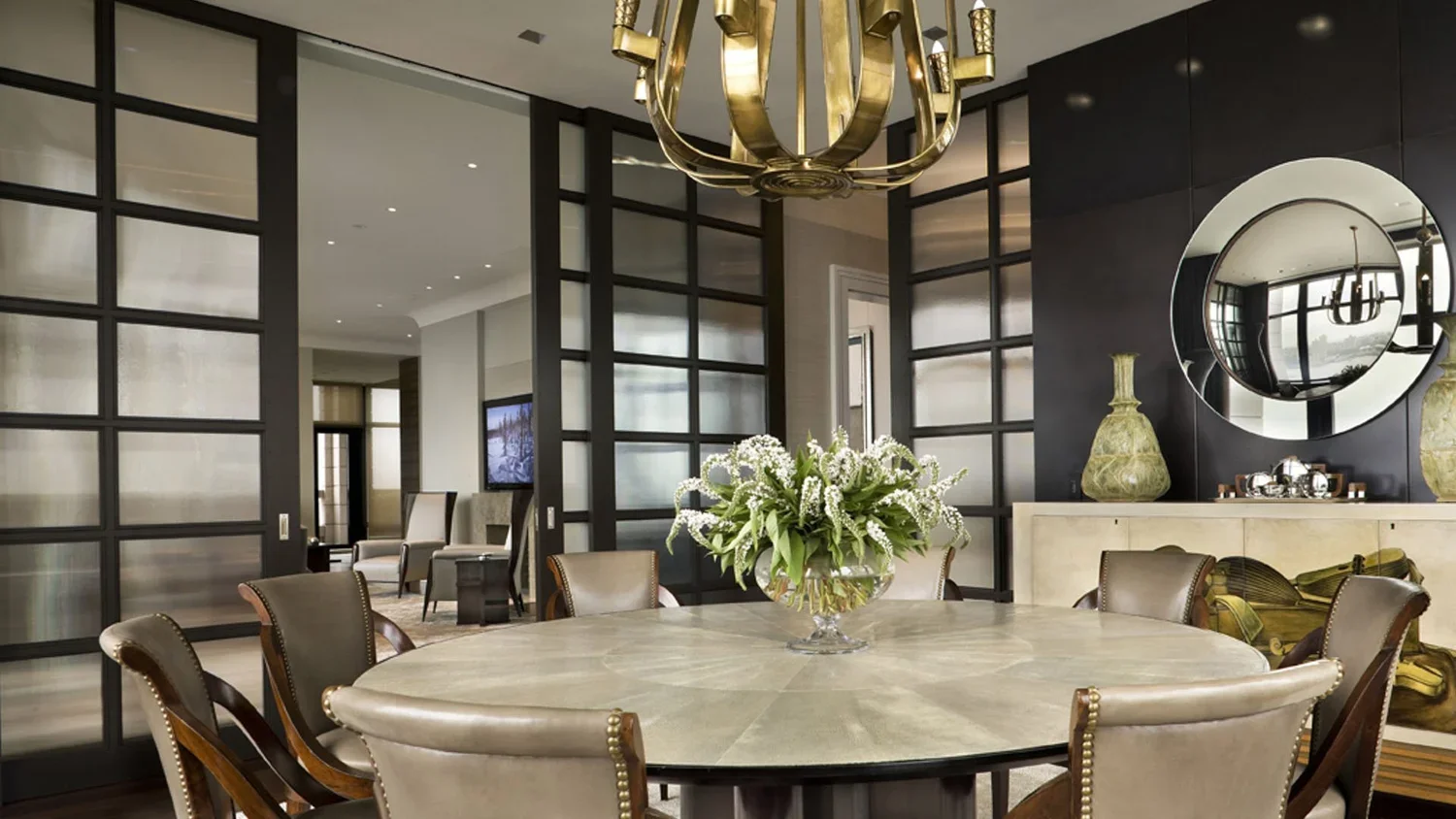Elegant dining room with round table, cream-colored chairs, a centerpiece with white flowers, a modern chandelier, and modern decor including vases and a round mirror.