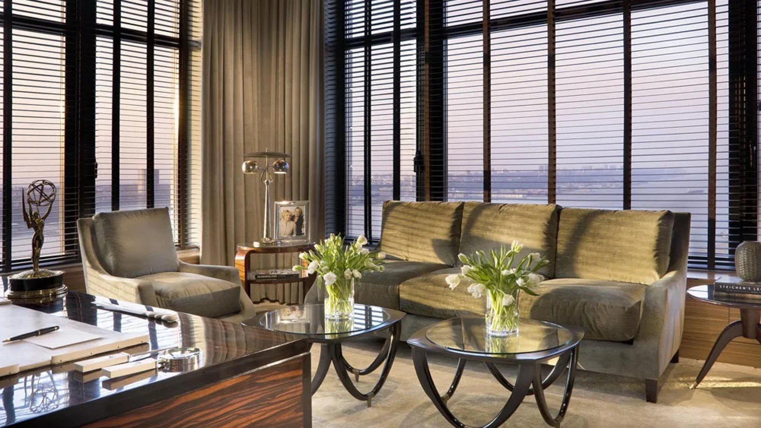 Modern living room with large windows, beige sofa, armchair, glass coffee tables with flowers, side tables with lamp and photo frame, and blinds.