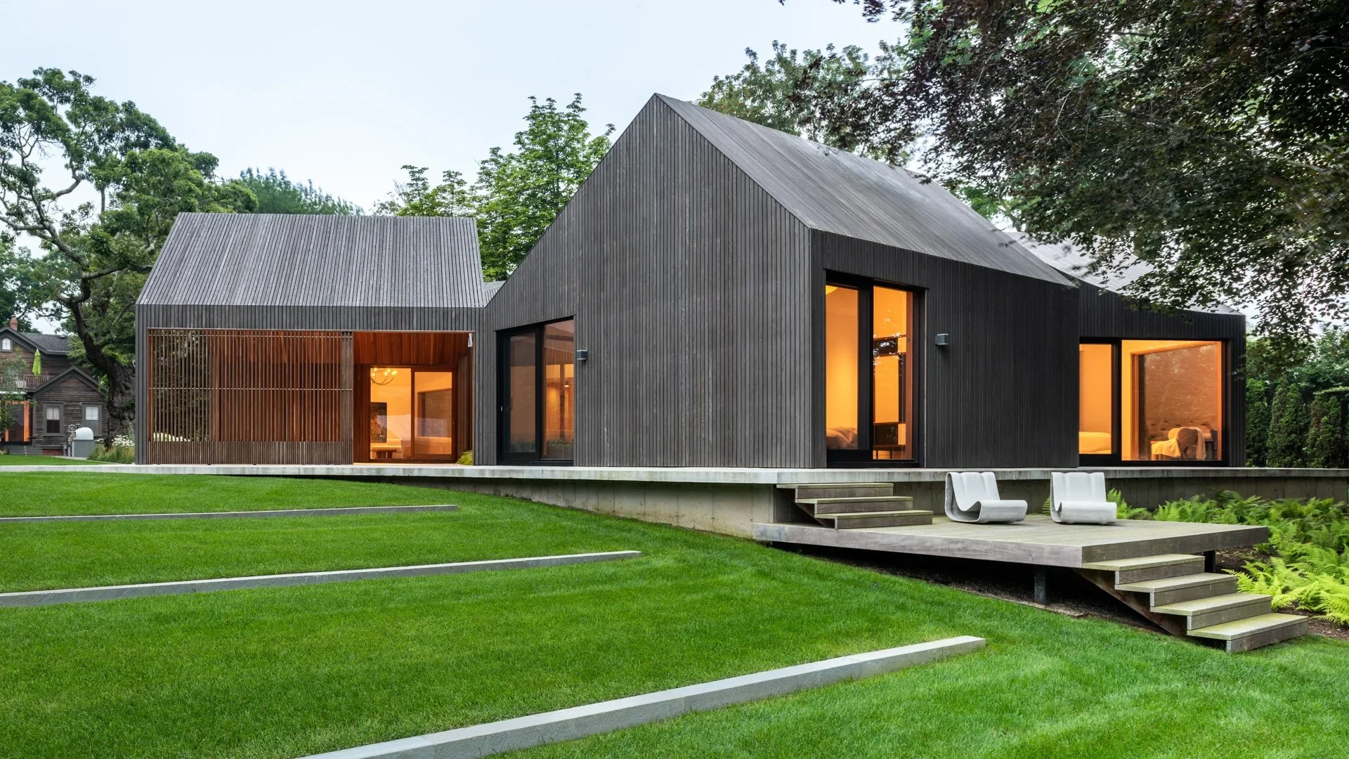 Modern house with black wooden exterior, large windows, and a raised wooden patio with white chairs, surrounded by green lawn and trees.