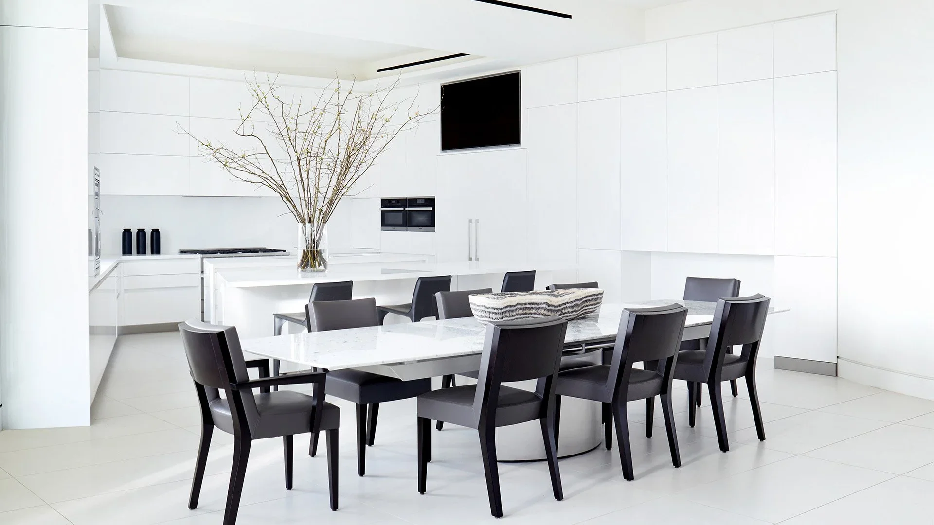 Modern white kitchen and dining area with black chairs around a white table, a tall vase with branches, and a black television on the wall.