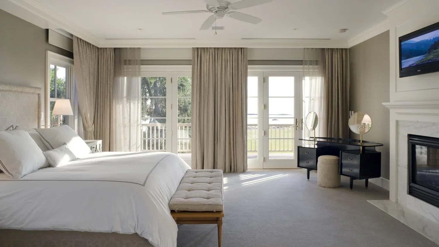 A bright, modern bedroom with large windows, cream curtains, a white bed with pillows, a beige bench at the foot of the bed, a black vanity with mirrors, a small beige ottoman, a TV mounted above a white fireplace, and light-colored walls and carpet.