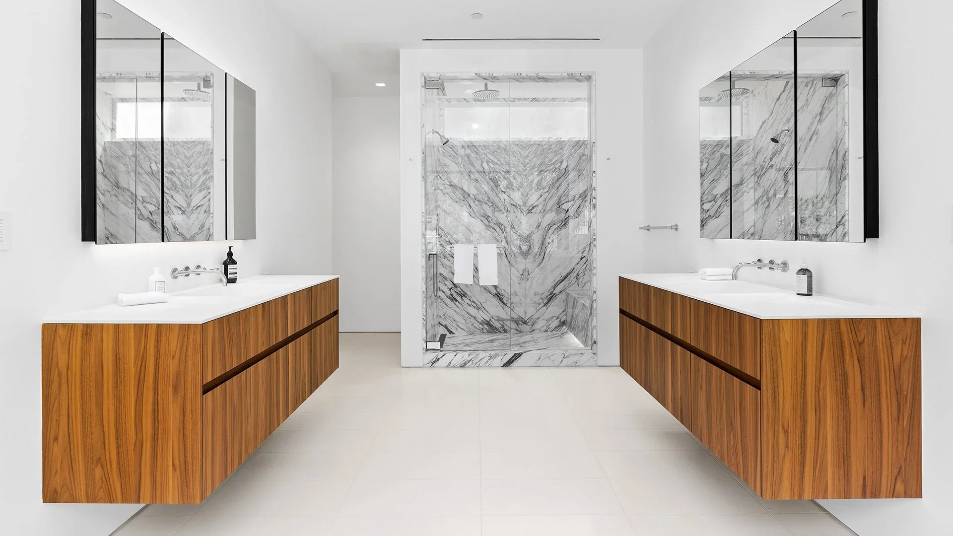 Modern bathroom with two wooden vanities, large mirrors, marble shower with glass door, and white walls and floor.