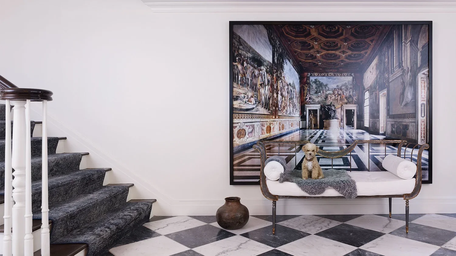A staircase with dark carpeted steps on the left, a white wall, and a decorative floor with black and white checkered tiles. A framed painting of an ornate interior hangs on the wall. In front of the painting, there's a vintage-style chaise lounge wi