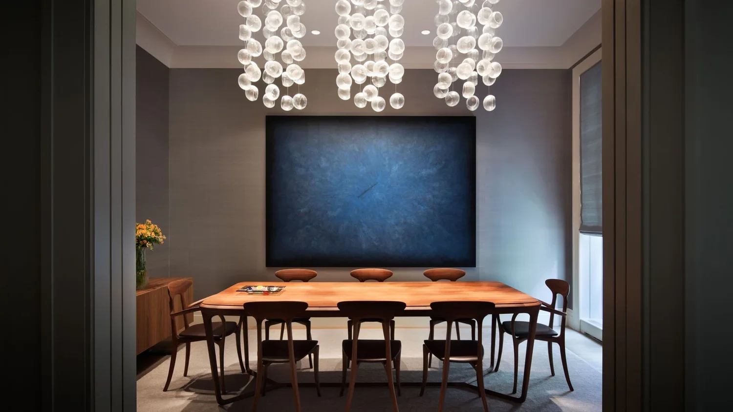 Modern dining room with a large wooden table surrounded by nine wooden chairs, a blue abstract wall art, a flower vase with yellow flowers, and a large window with blinds. A chandelier with multiple glass orbs hangs from the ceiling.