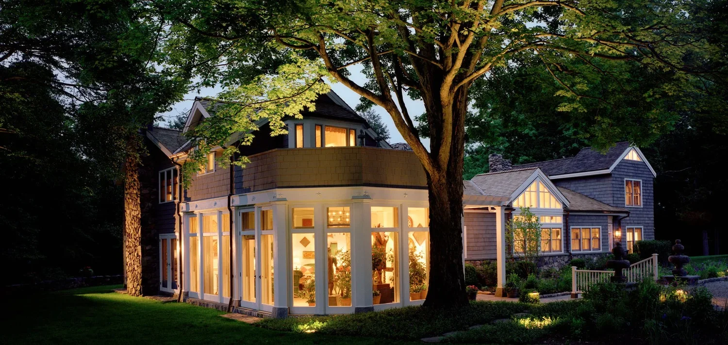 A house with large windows illuminated from inside, surrounded by trees and a well-maintained yard with garden lights at dusk.