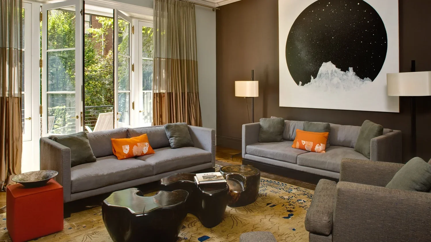 Living room with large windows and garden view, featuring two gray sofas with orange and gray pillows, a dark wood coffee table, a patterned area rug, a floor lamp, and abstract wall art of a starry night above a mountain.