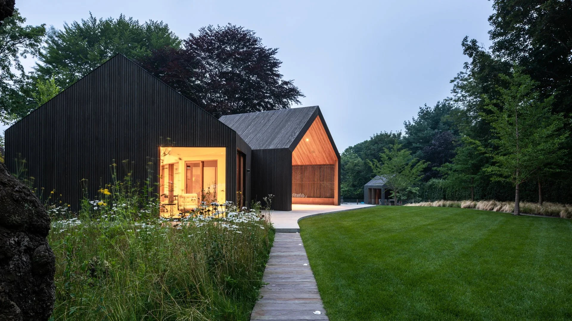 Modern black and wood house with large windows, surrounded by a lush green lawn and trees.