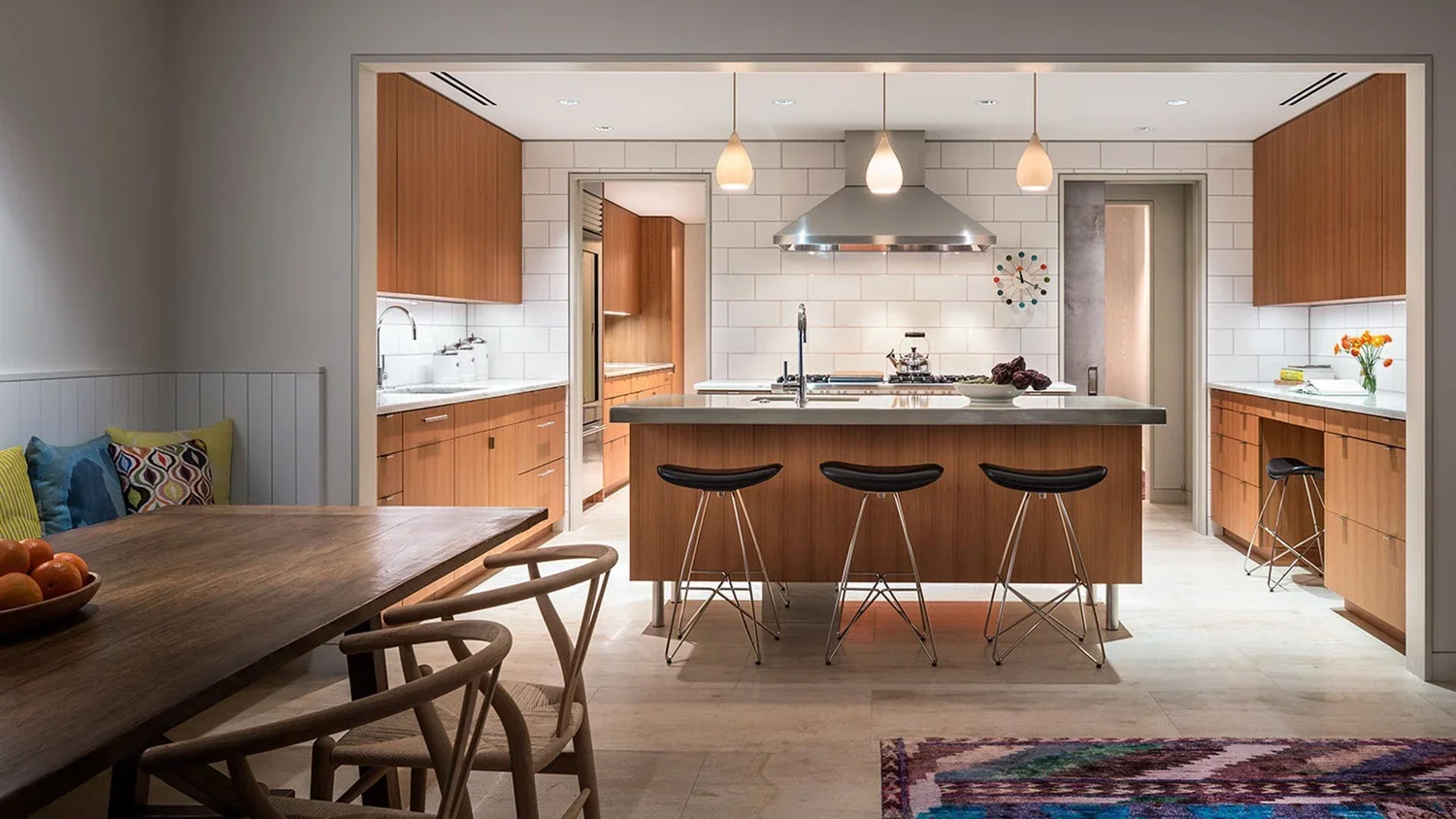 A modern kitchen with a central island, wooden cabinets, white tile backsplash, and hanging pendant lights. There are three black bar stools at the island. To the left, a dining table with chairs and a bowl of fruit is visible, while to the right, a 