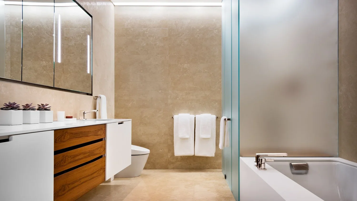 Modern bathroom with beige stone walls and floor, white vanity with wooden drawers, large mirror, potted plants, white towels, toilet, and bathtub with glass partition.