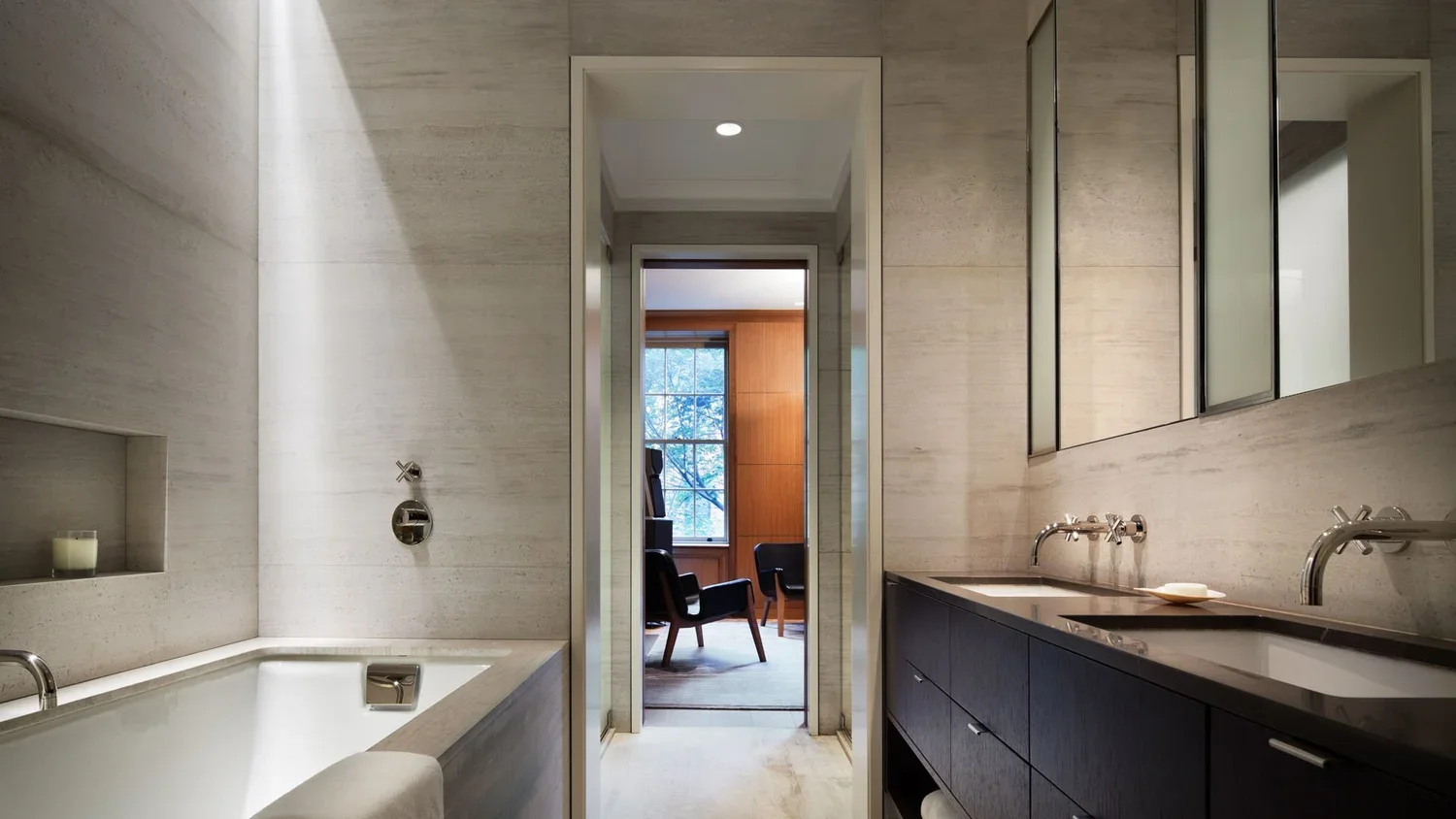 Modern bathroom with beige tiled walls, a bathtub on the left, and a dark-colored double sink vanity on the right. Through a doorway, a room with a large window and two chairs can be seen.
