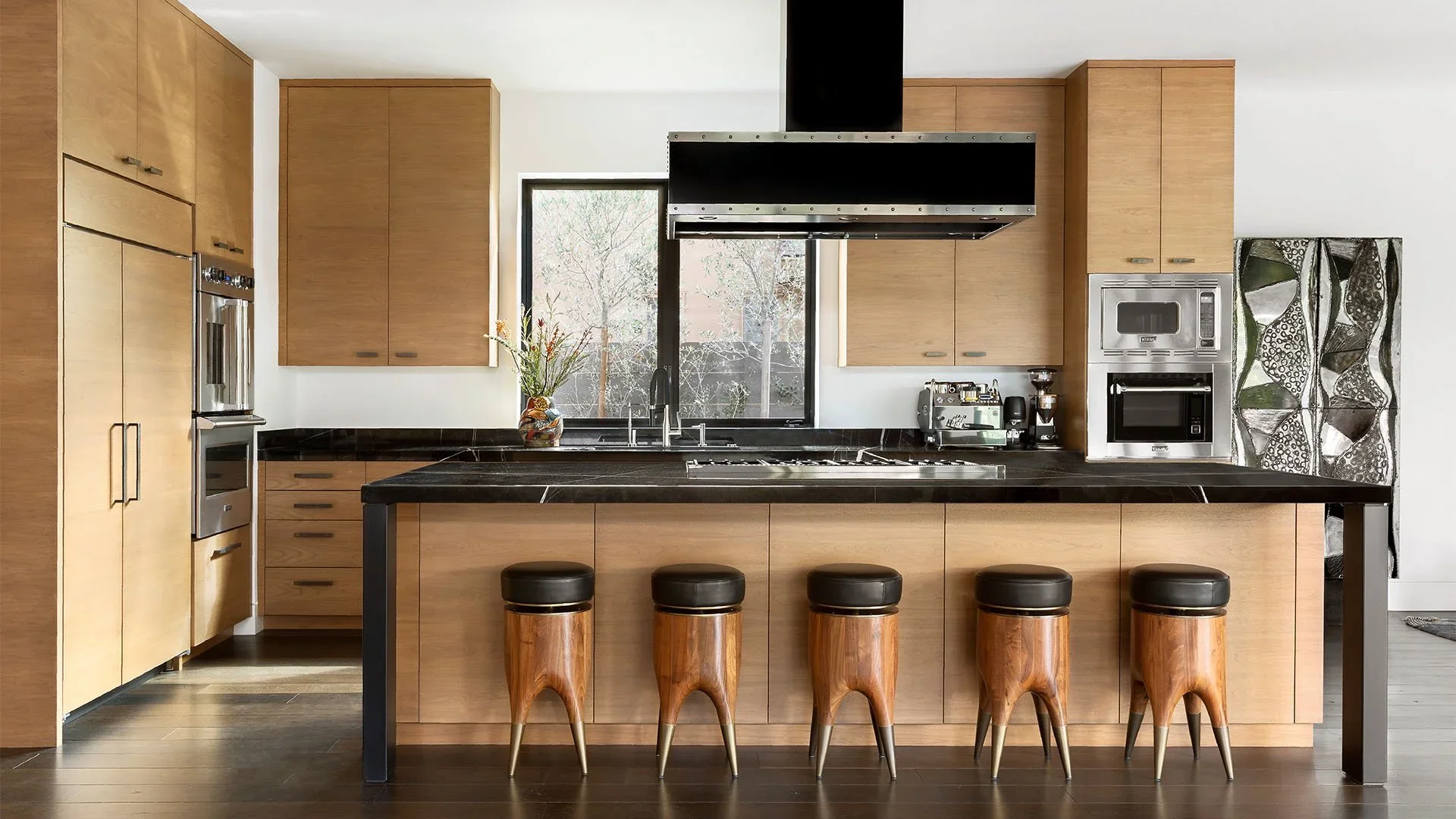 Modern kitchen with wooden cabinets, black countertop, and stylish bar stools at the island, featuring a window behind the sink and contemporary appliances.