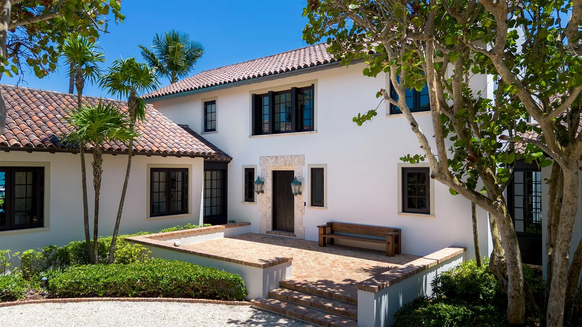White house with black window frames, terracotta tiled roof, and a small courtyard with steps, brick paving, a bench, and trees