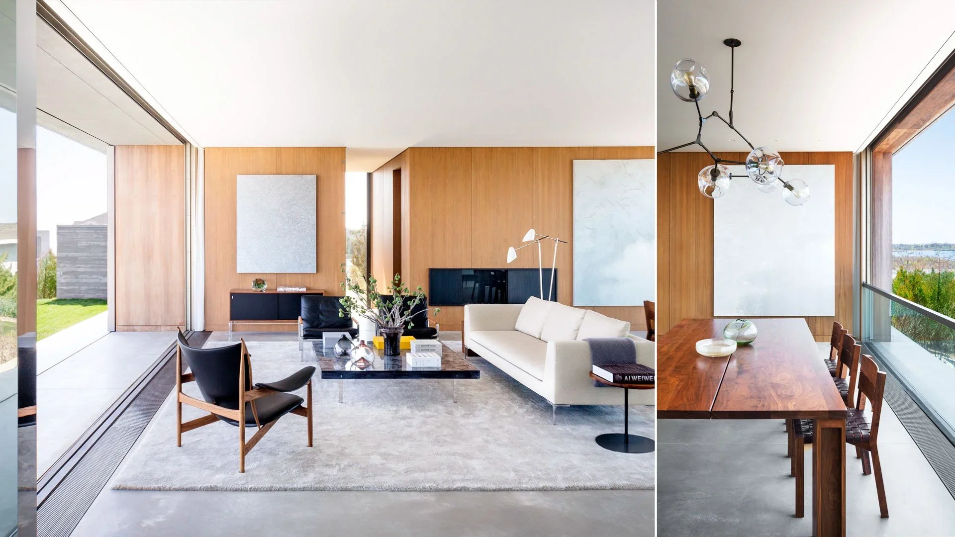 Living room with large glass windows, wooden walls, modern furniture including a white sofa, black armchairs, a marble coffee table, and abstract art on the walls. Adjacent dining area has a wooden table, chairs, and a modern chandelier.