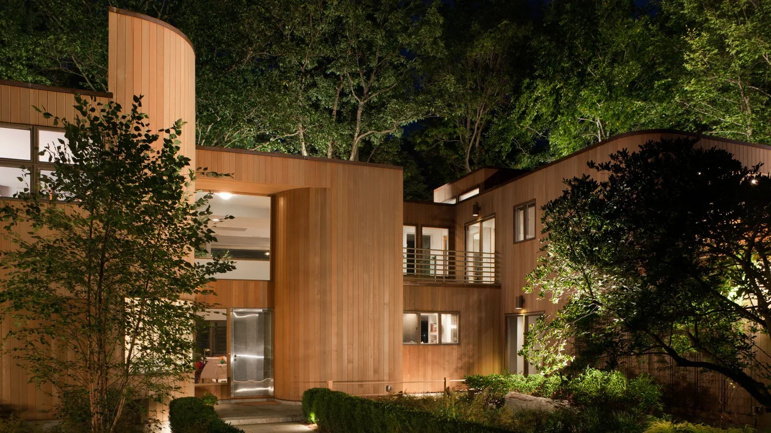 Modern wooden house at night with trees and greenery surrounding it, illuminated from inside.