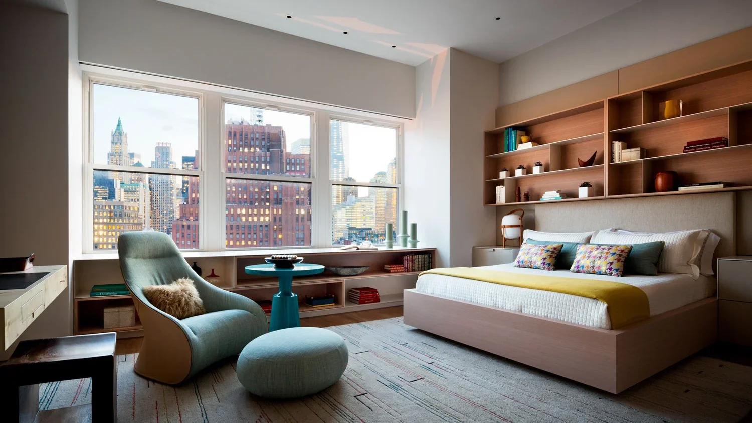 Modern bedroom with large windows showcasing a city skyline. The room has a bed with colorful pillows and a yellow blanket, a light blue armchair with a matching ottoman, and wooden shelving with decorative items and books.